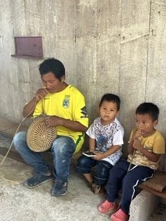 An older boy in a yellow shirt with a Brazil logo spinning a large wooden top while two young children watch. They are sitting against a rough concrete wall, with a small purple shelf in the background. In the Amazon, Colombia.