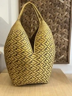Yellow and black patterned woven bag with a slot opening, placed on a wooden surface in front of a decorative lattice background. Known as Cuatro Tetas baskets in Colombia.