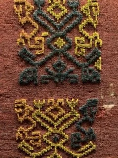 Close-up of a woven textile with intricate geometric patterns in yellow, blue, and brown on a reddish-brown background.