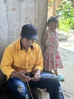 A man in a yellow shirt and black cap sitting outdoors, using his phone, with a girl in a floral dress standing nearby, in the Amazon, Colombia.