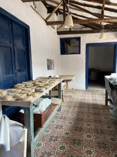 Interior of a restaurant or cafe with white walls, blue window and door frames, patterned tile floor, wooden ceiling beams, and a long table set with plates and napkins.