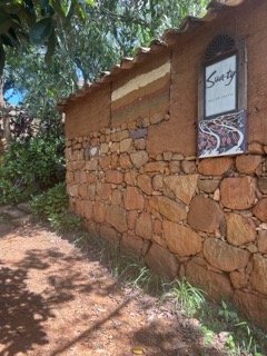 A rustic stone wall with a window displaying the word "San Jose" on a building surrounded by trees and bushes.