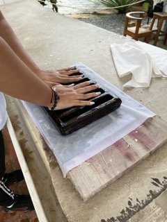 Person pressing yeast dough with hands on a black baking stone, covered with a white cloth, on a wooden table in an indoor setting.