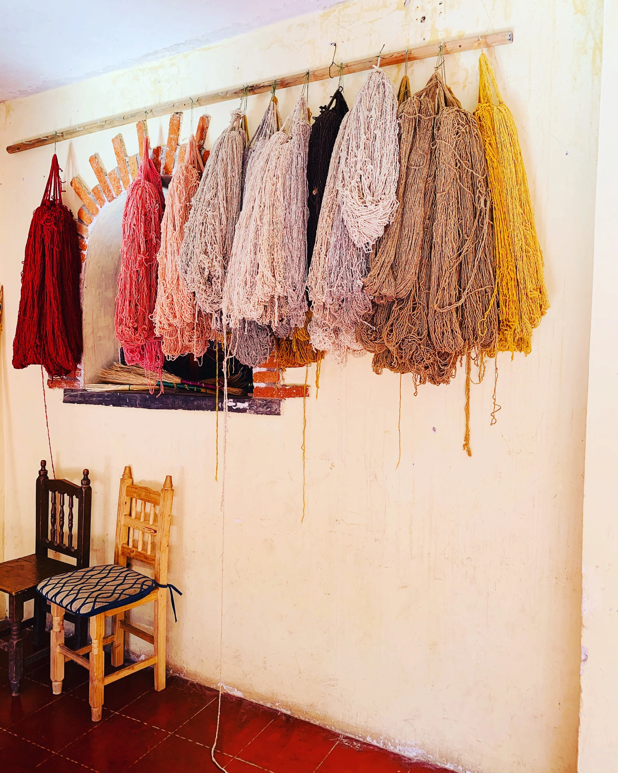 Colorful yarn bundles hanging on a wooden rack above two wooden chairs in a cozy room with a small window in Oaxaca.