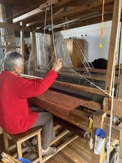 An elderly woman in a red sweater sits on a wooden chair, weaving fabric on a traditional loom in a workshop or craft room, in Bogota Colombia.