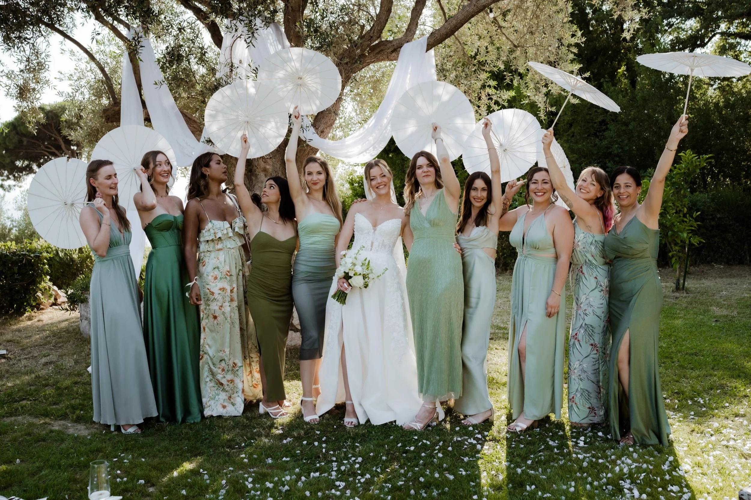 A bride in a white dress holding a bouquet of white flowers standing next to bridesmaids in green dresses holding white parasols in an outdoor garden setting with trees and greenery.
