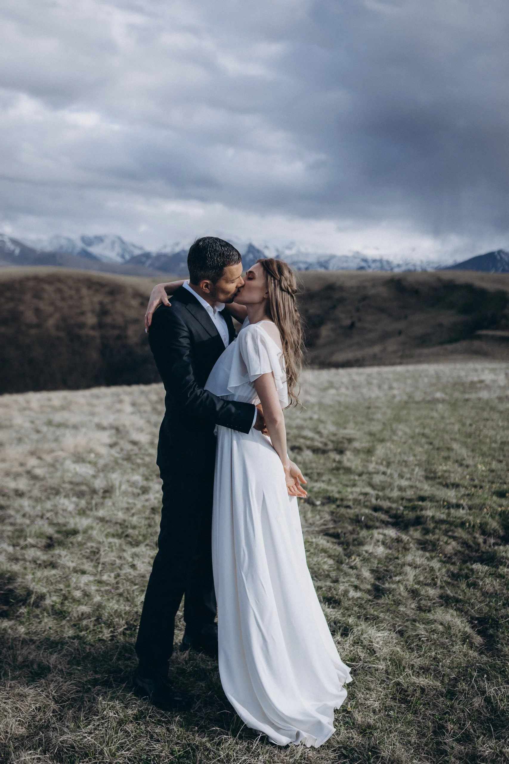 A man and woman are kissing in an open field during daytime, with mountains and a cloudy sky in the background. The woman is wearing a white dress, and the man is in a black suit.