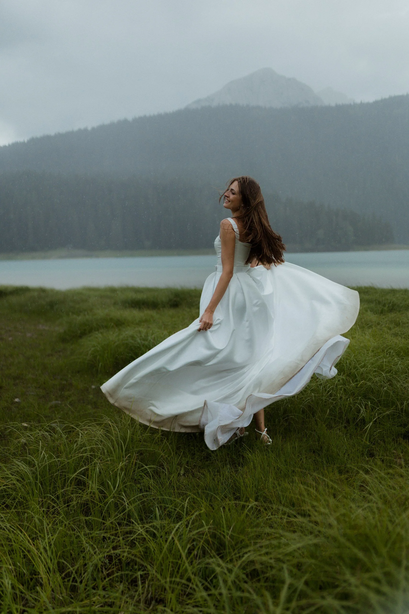 A woman in a flowing white dress smiling and spinning in a grassy field near a body of water with mountains in the background during overcast weather.