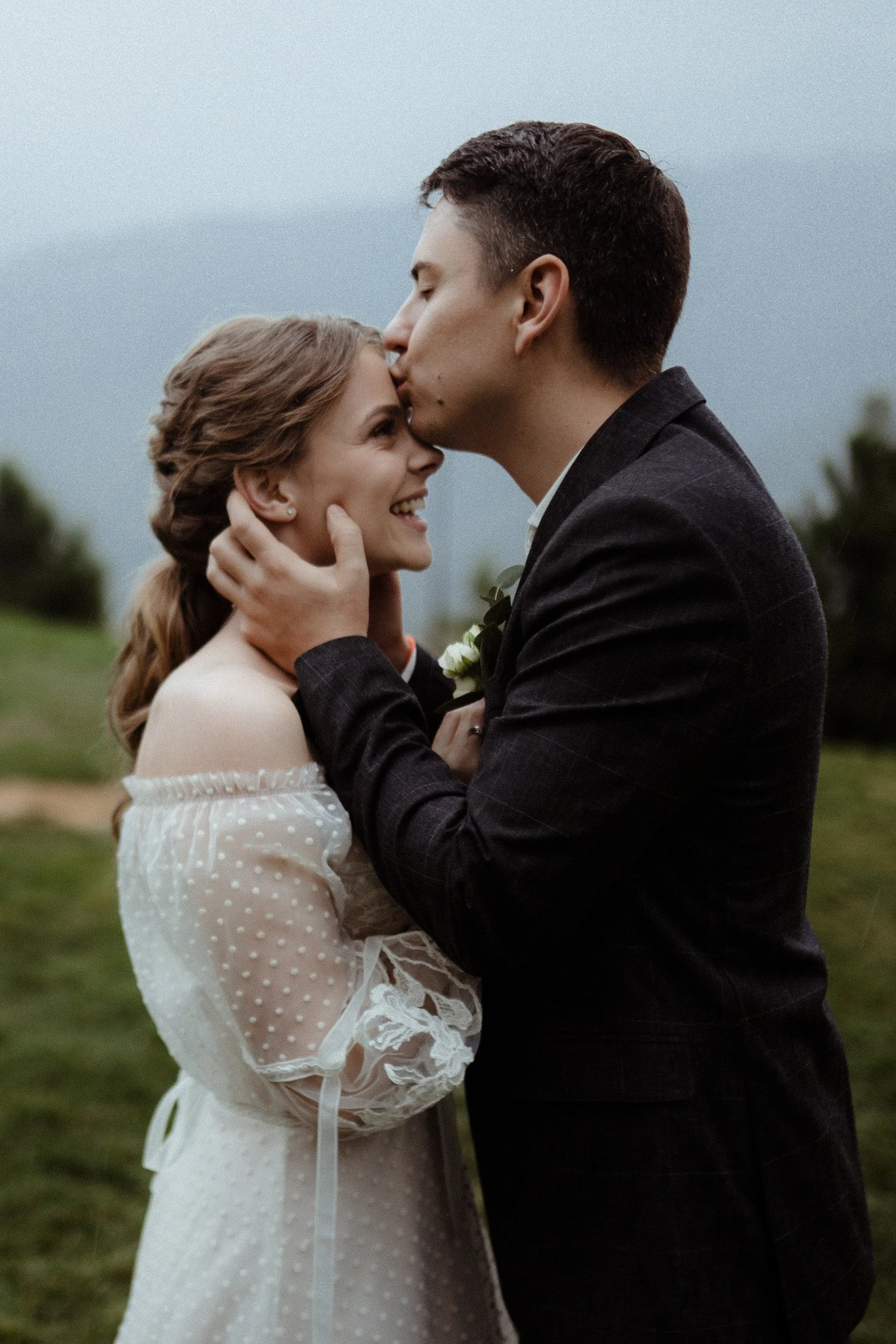 A bride and groom share a kiss outdoors, with the groom kissing the bride on the forehead, and the bride smiling while holding the man's face. The groom is wearing a dark suit, and the bride is wearing a white wedding dress with lace details.