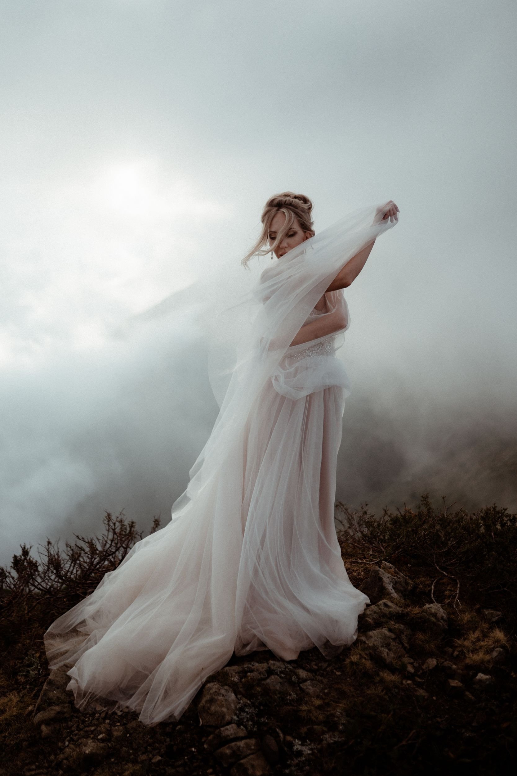 A woman in a flowing white dress stands outdoors on rocky terrain, with dark clouds overhead and a cloudy sky in the background.