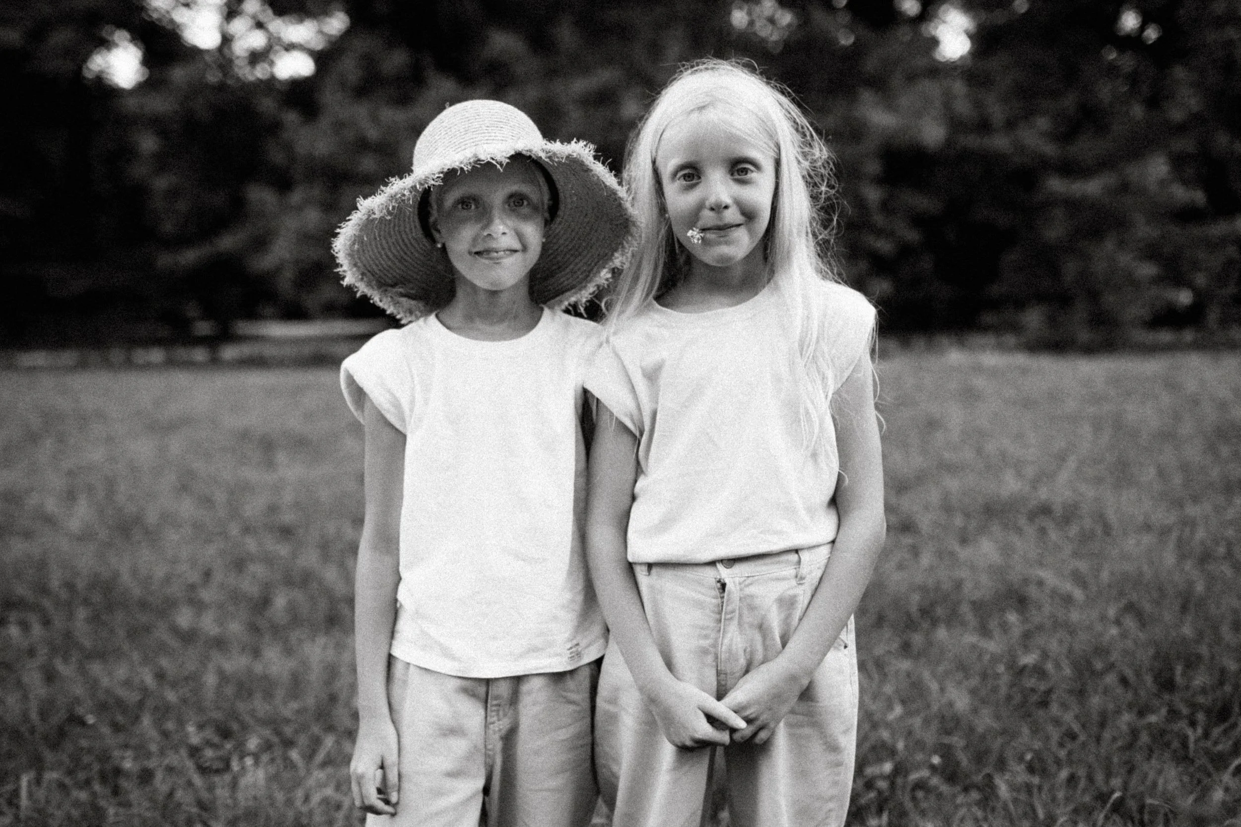 Two young girls standing together outdoors, one wearing a large sunhat and the other with long hair, both dressed in light-colored shirts and pants, with a blurred natural background.