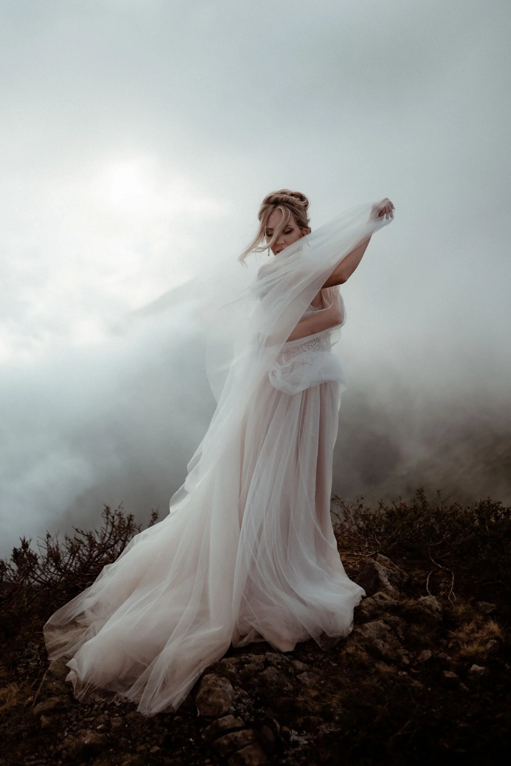 A woman dressed in a flowing white gown stands outdoors on a rocky terrain, with an overcast sky in the background.