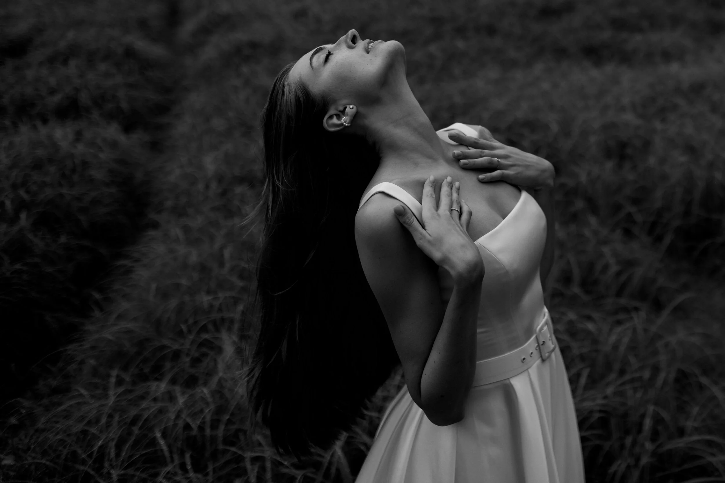 Black and white photograph of a woman with long hair, her head tilted back, eyes closed, with her hands resting on her neck and chest, standing outdoors on grass.