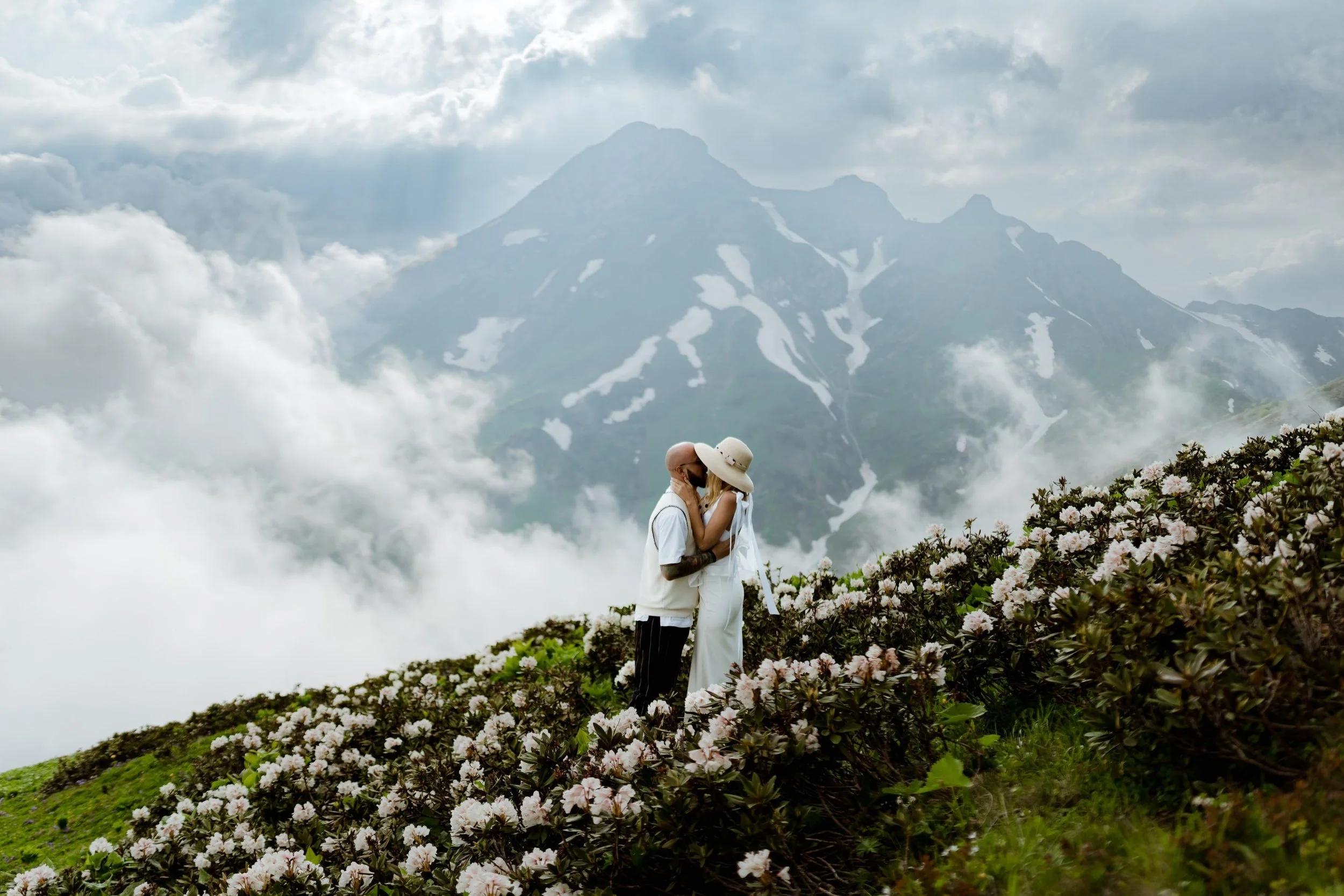 A couple embracing on a mountain trail covered with blooming pink and white flowers, with foggy mountain peaks and cloudy sky in the background.