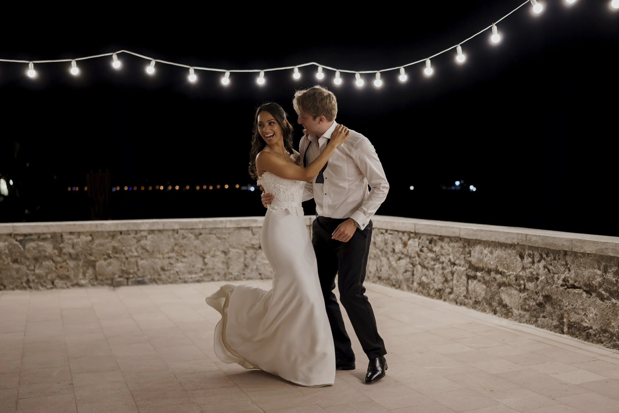 A bride and groom dancing outdoors at night under string lights, with a dark sky and city lights in the background.