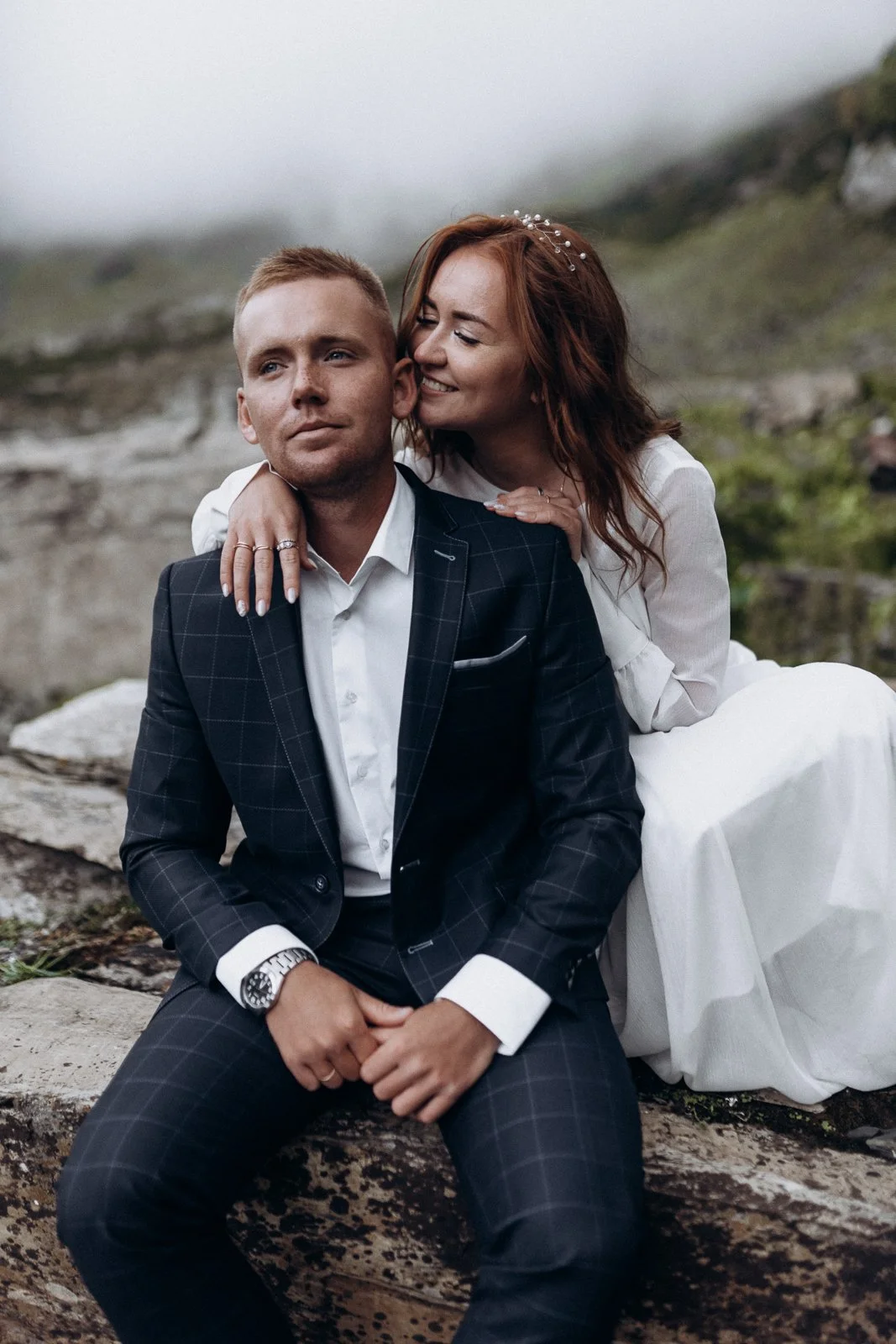 A couple sitting outdoors, the woman with red hair and white dress whispering to the man in a checkered suit, on a rocky landscape with a misty background.