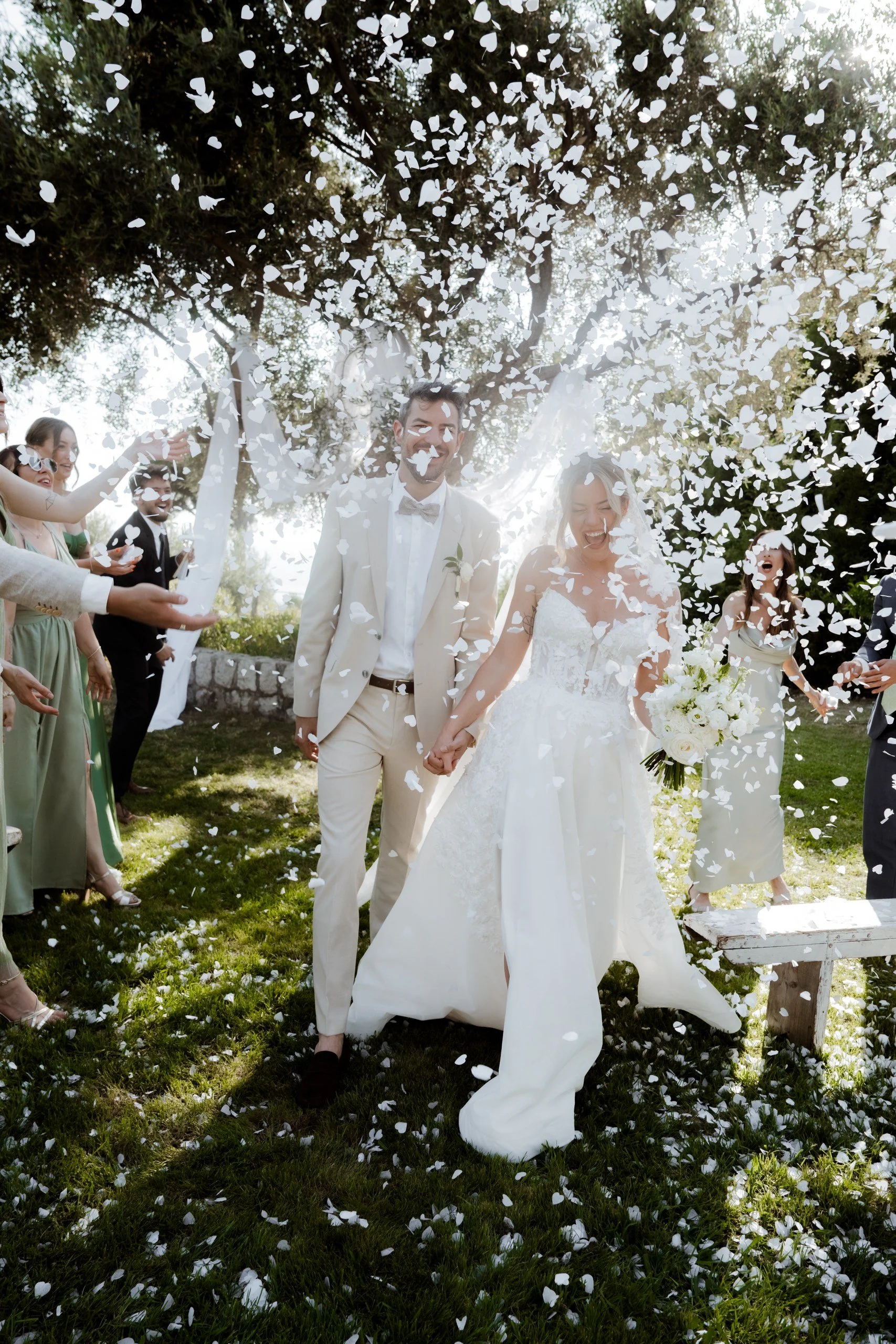 Bride and groom holding hands smiling as they walk through falling white flower petals outdoors during wedding celebration with friends.