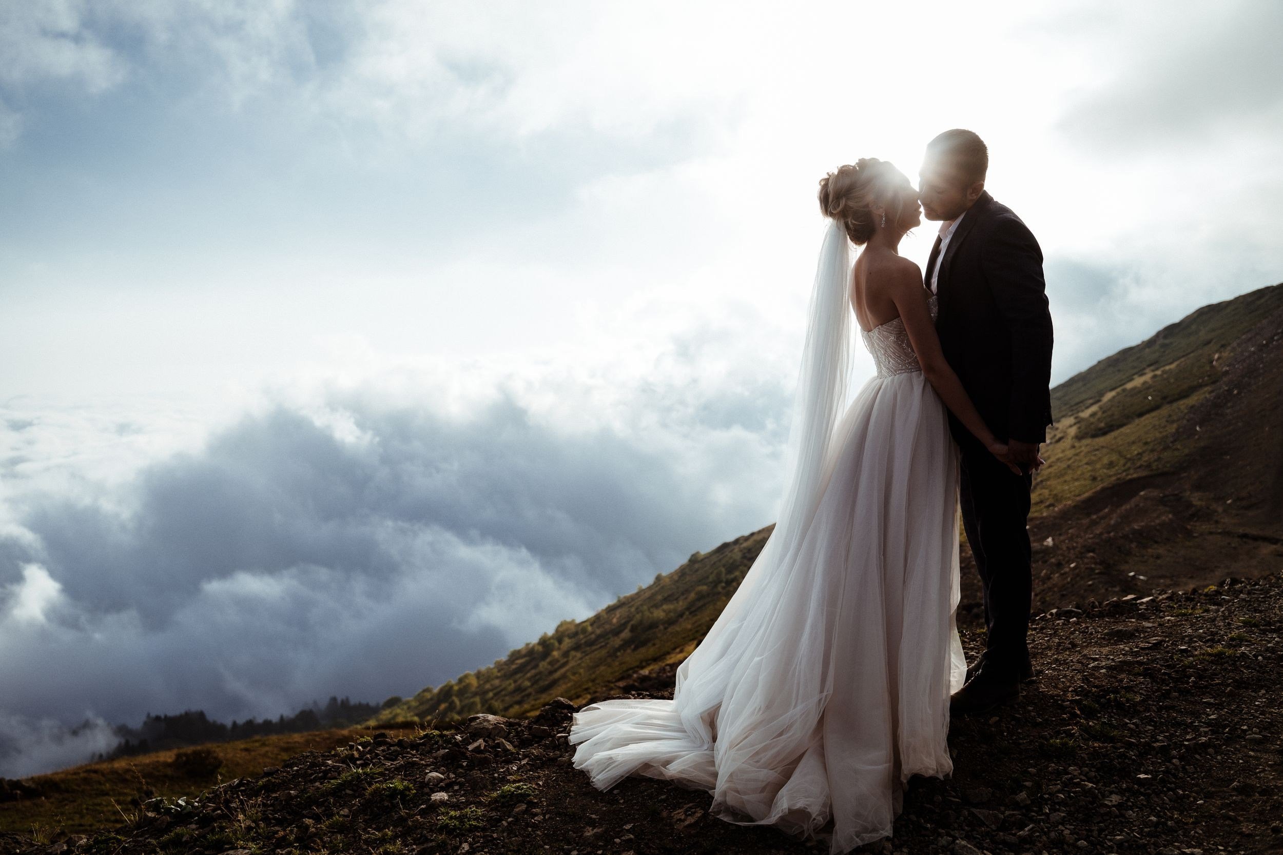 A bride and groom sharing a kiss outdoors against a cloudy sky and mountain landscape, with the bride wearing a white wedding dress and the groom in a dark suit.