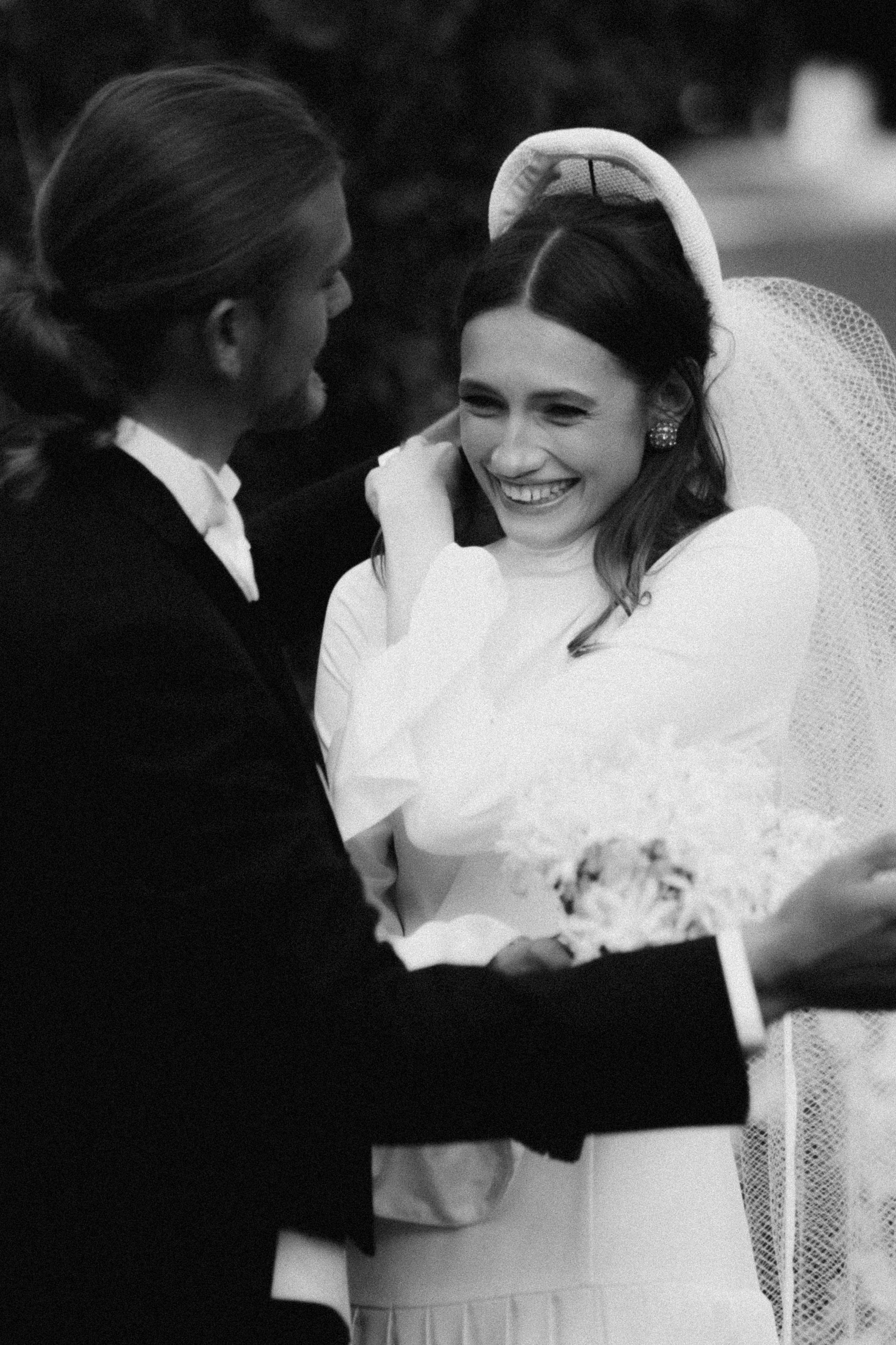 A black and white photo of a wedding scene with a man and woman. The man, with long hair tied back, is smiling and looking at the woman. The woman, wearing a bridal veil, is smiling and looking at the man, with her hand near her face. She is wearing earrings and a white dress, holding a bouquet.
