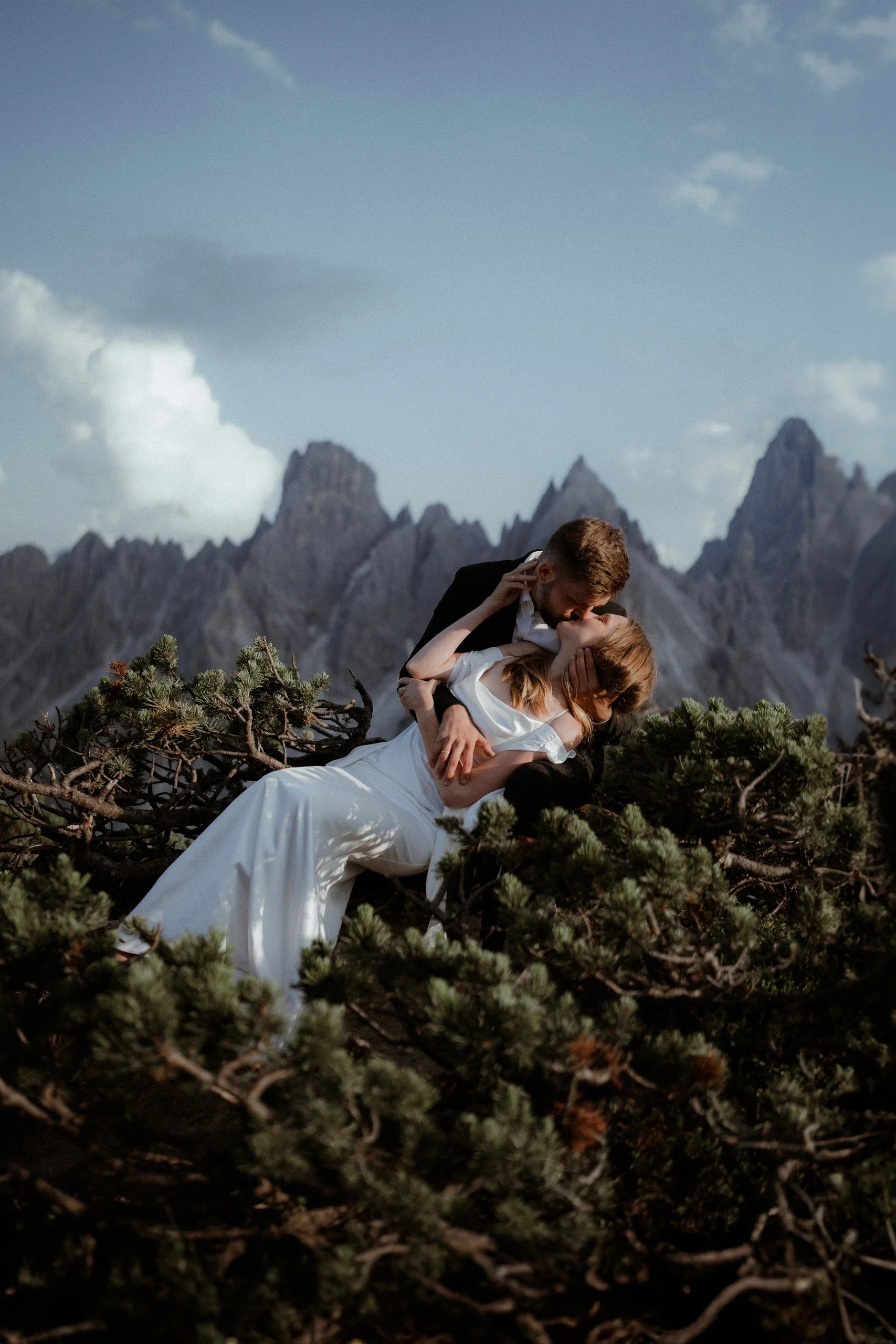 A couple dressed in wedding attire sharing a kiss outdoors amidst plants with mountains in the background.