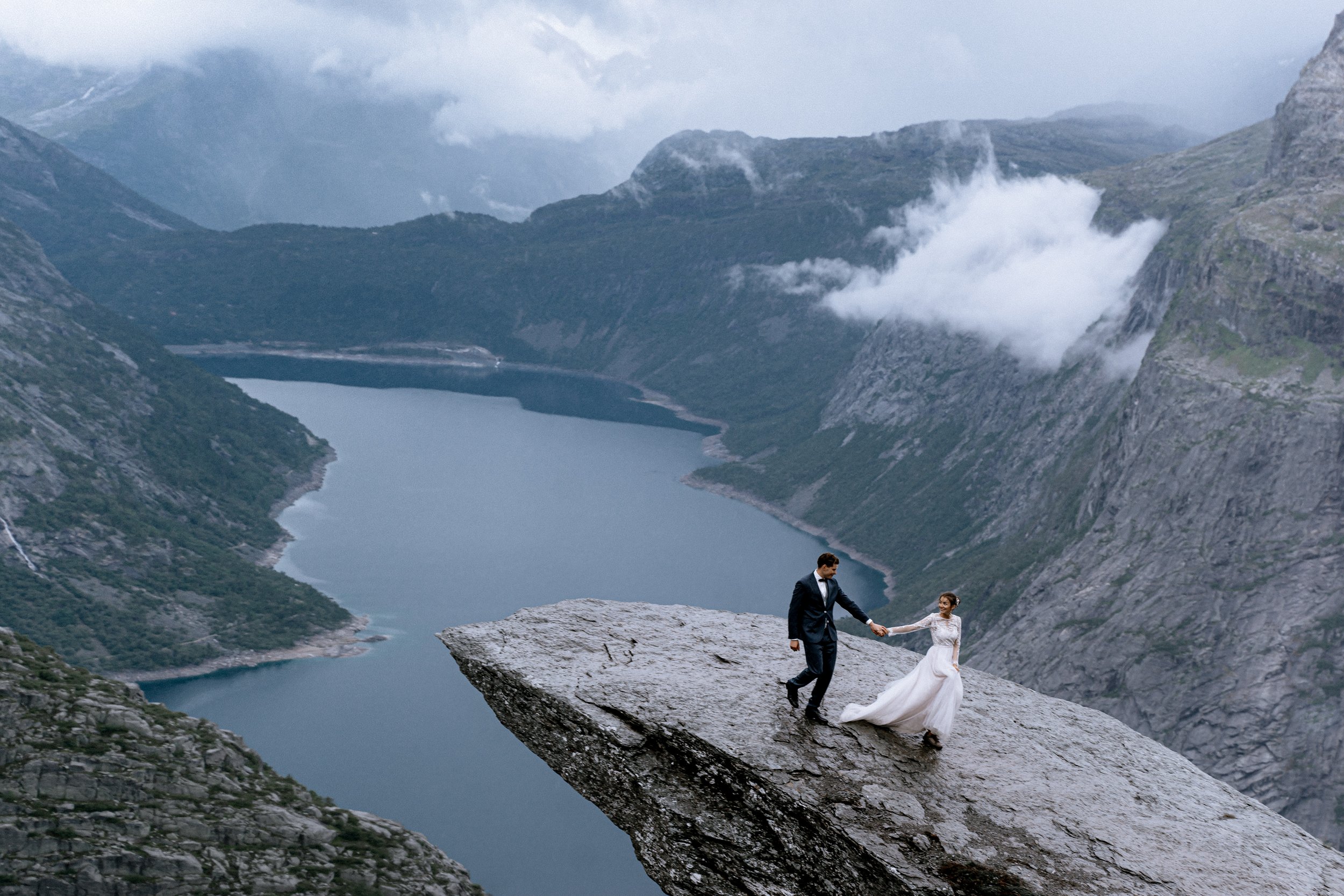 A bride and groom holding hands on a large rock ledge with a scenic mountain lake and cloudy sky in the background.