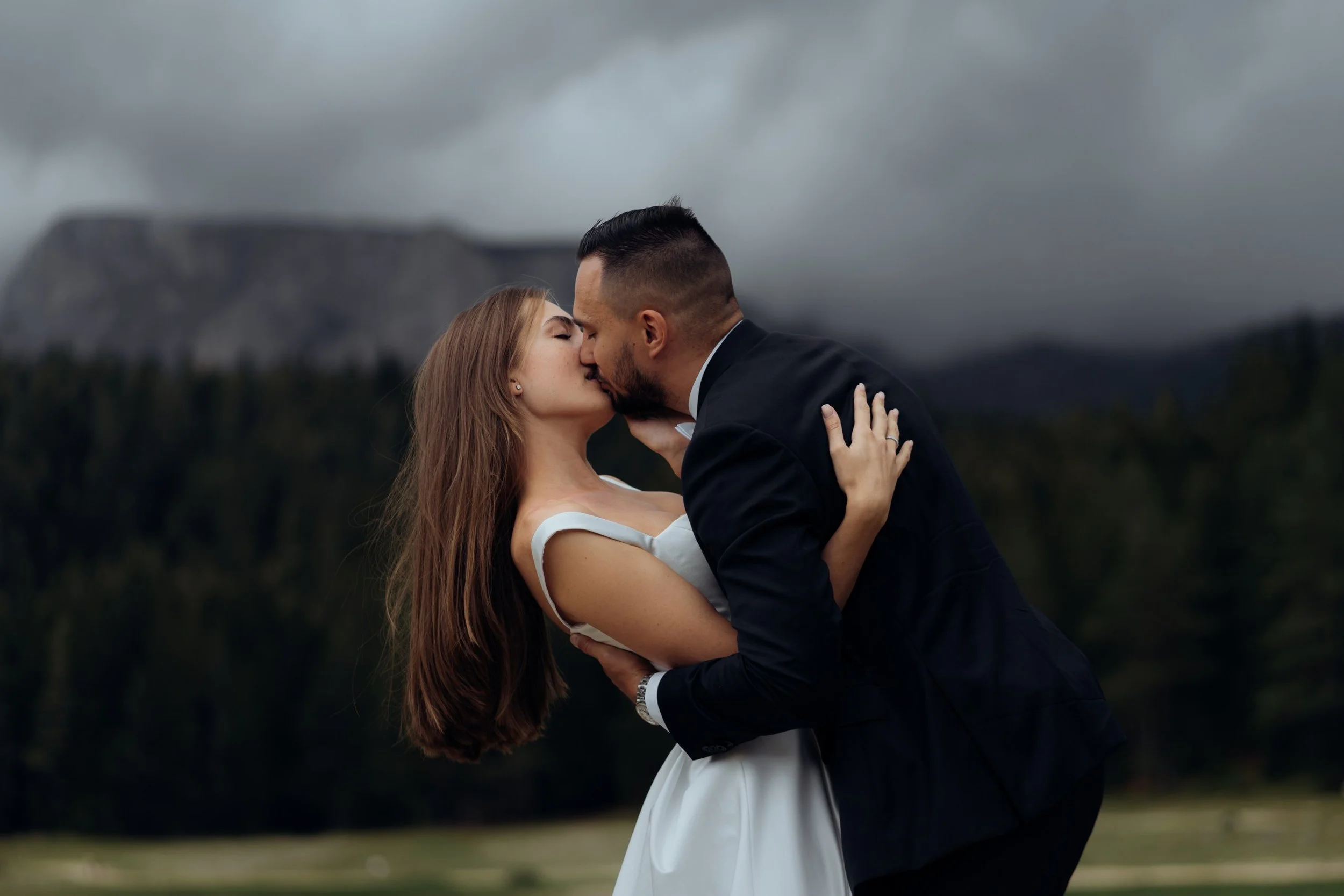 A couple dressed in wedding attire sharing a kiss outdoors on a cloudy day, with mountains and forests in the background.