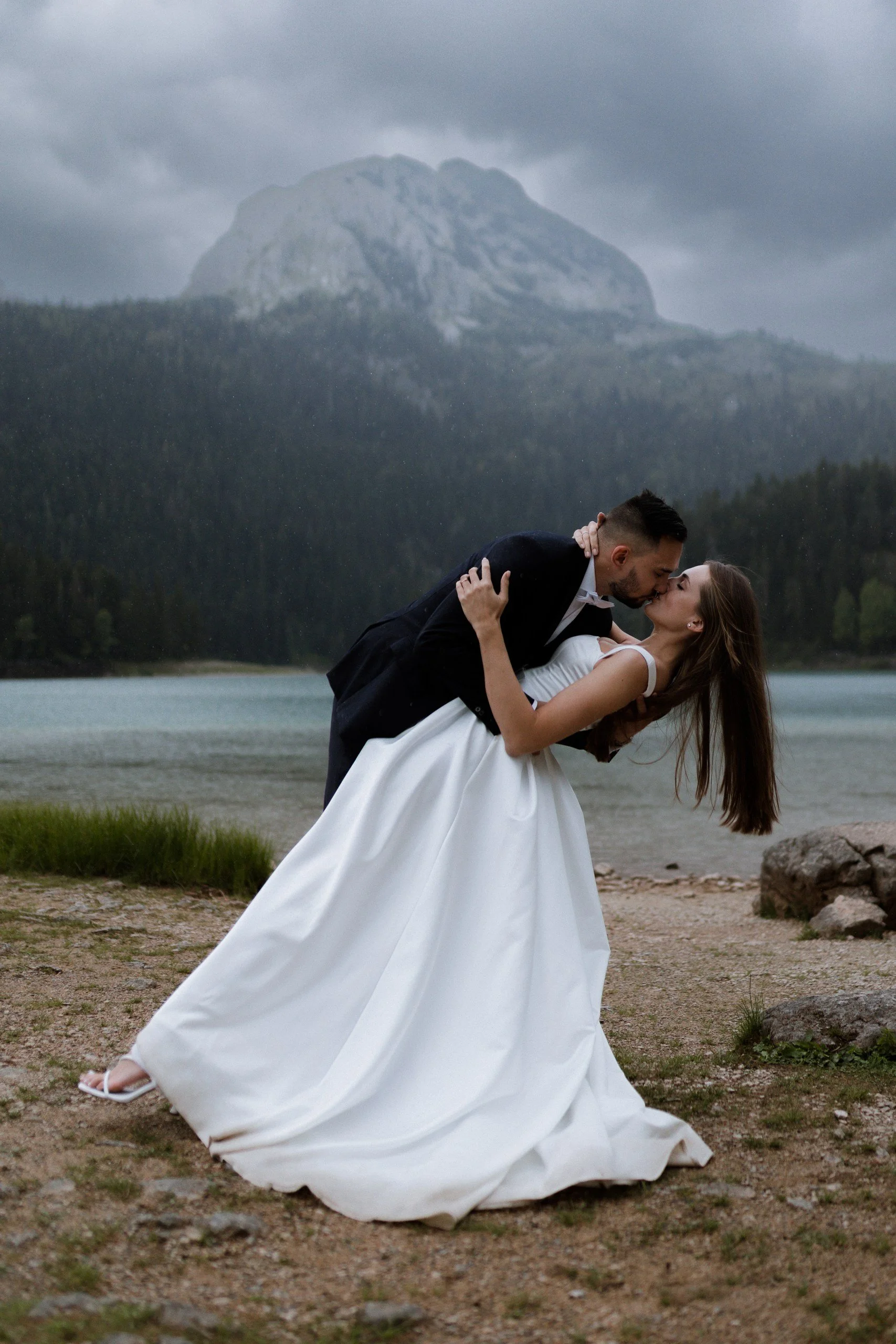 A couple, with the man in a tuxedo and the woman in a white wedding gown, sharing a kiss outdoors near a lake, with mountains and cloudy sky in the background.