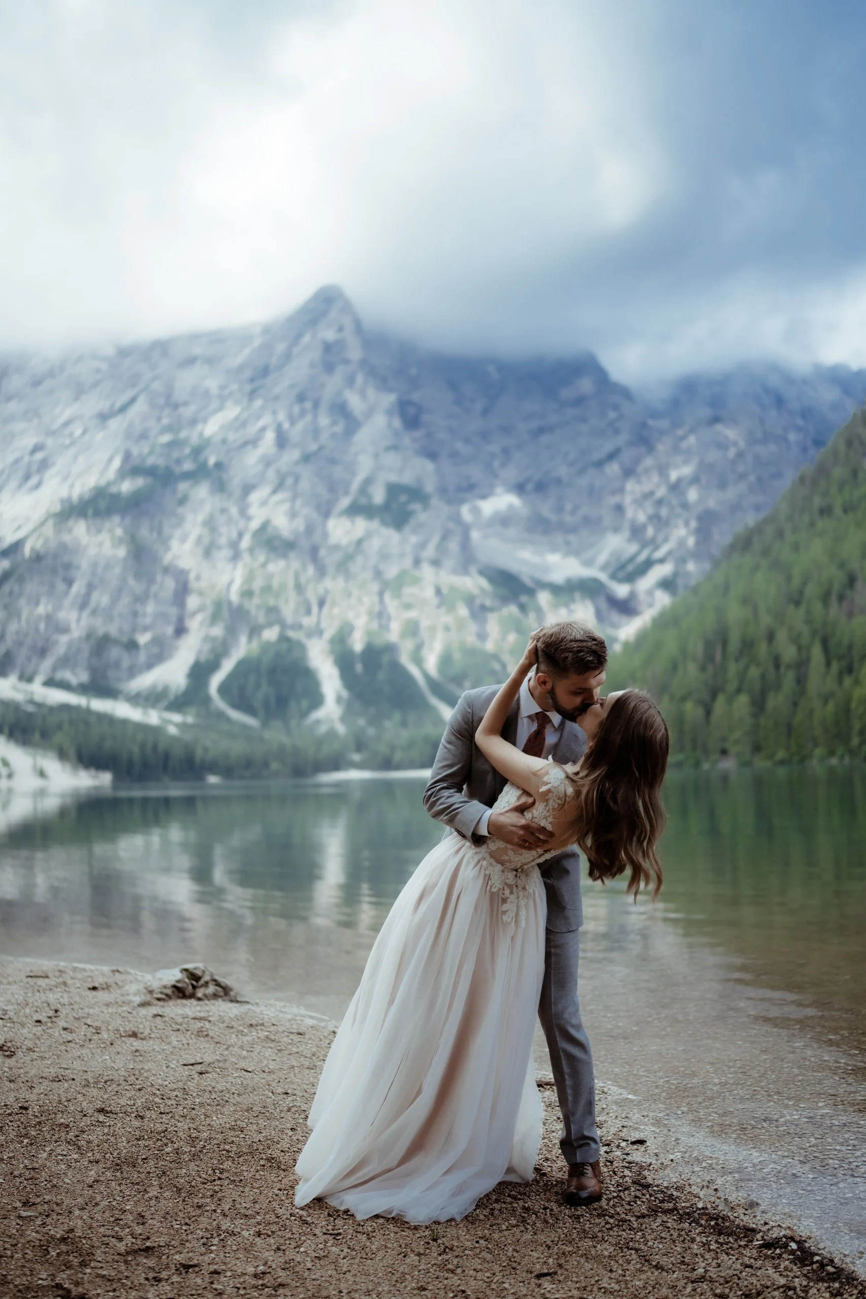 A couple in wedding attire sharing a kiss by a lake with mountains in the background.
