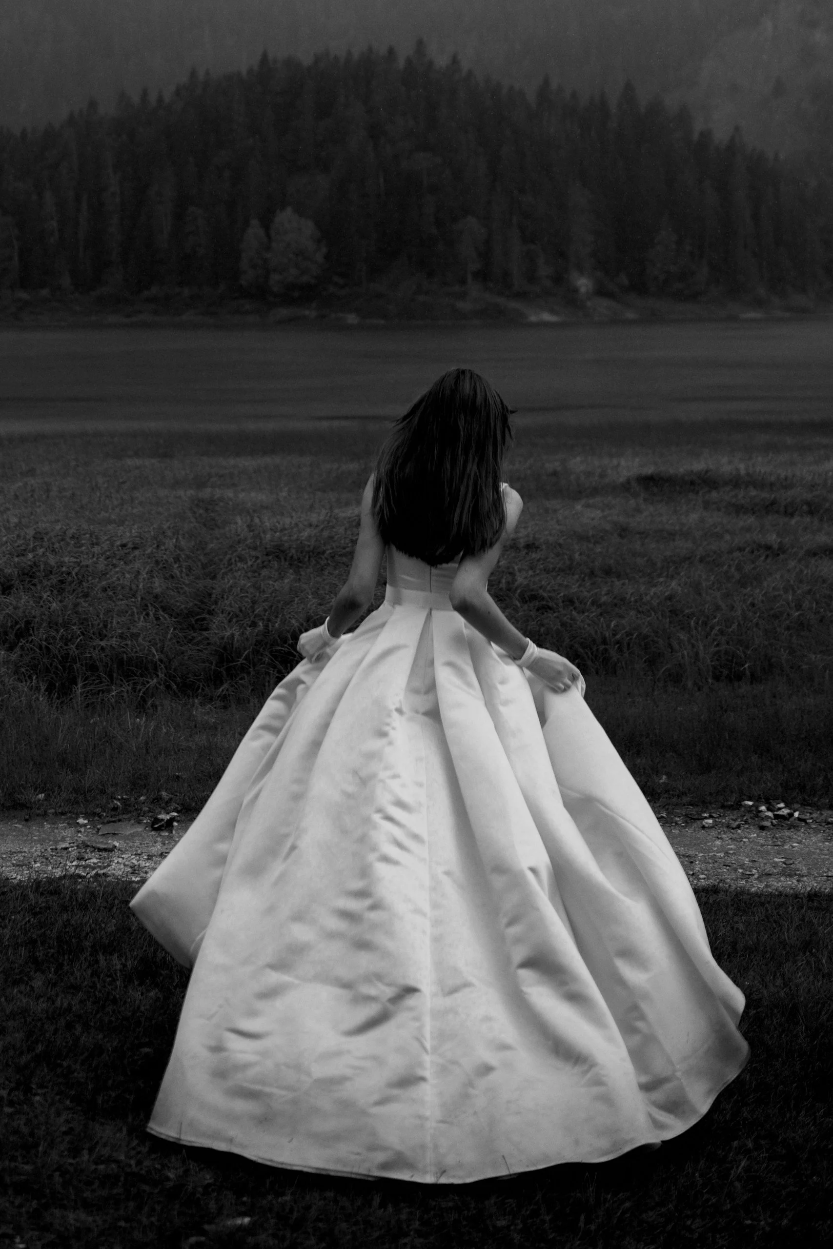 A woman in a white wedding dress sitting on the grass facing away, with dark trees and a body of water in the background.