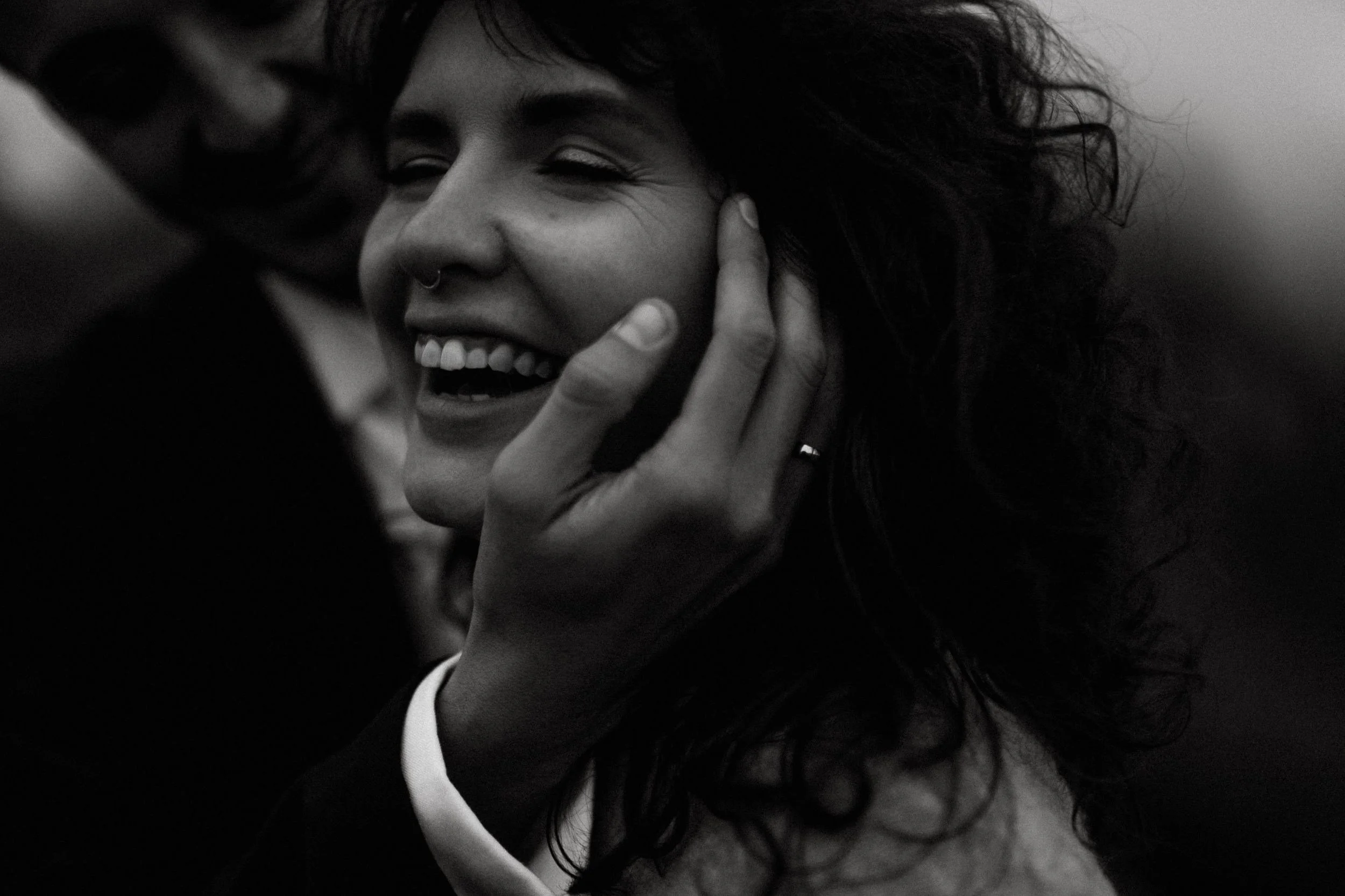 A person with curly hair smiling and touching their face, in black and white.