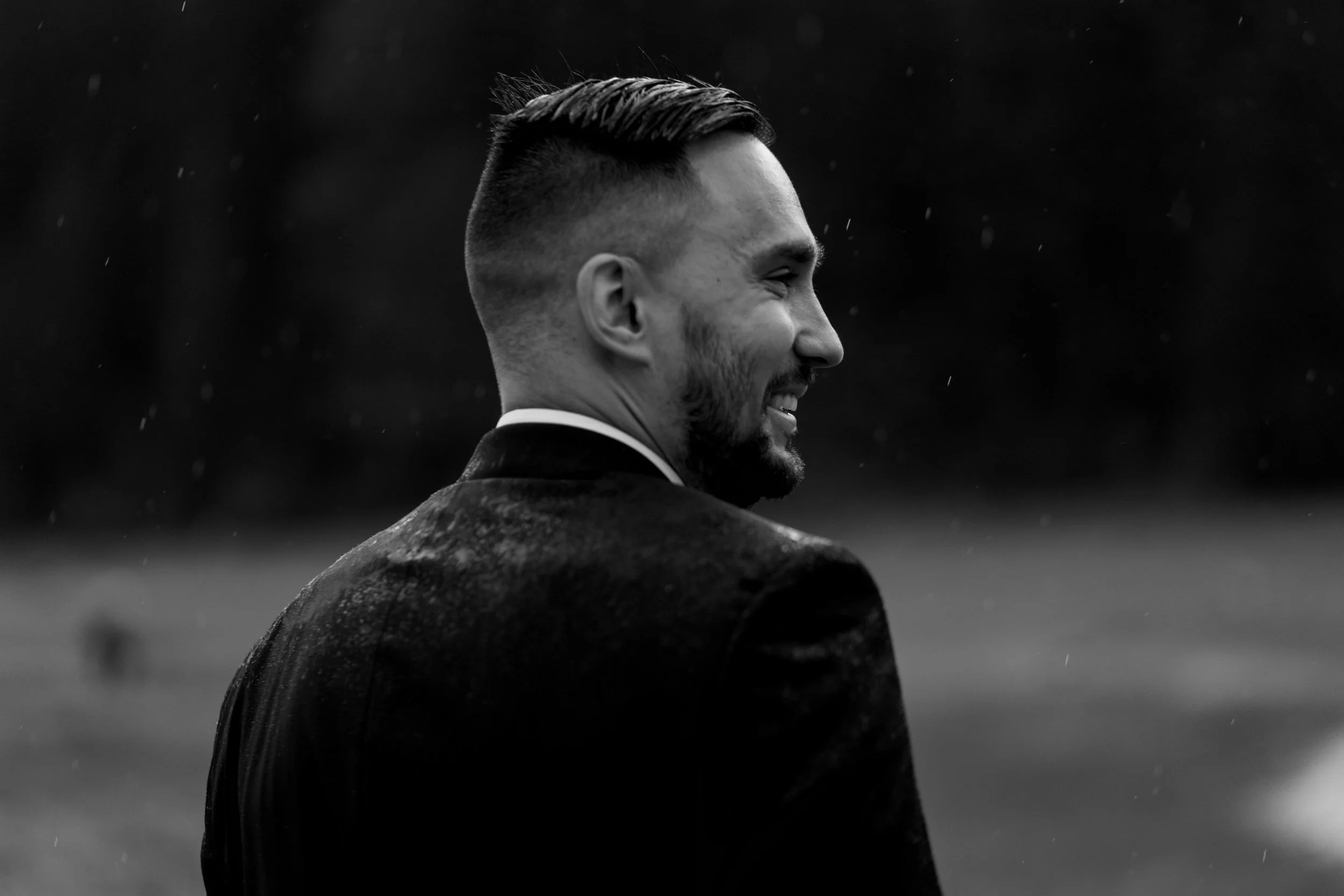 A black-and-white photo of a man smiling, facing right, with short dark hair and a beard, wearing a suit jacket, in an outdoor setting with blurred background.