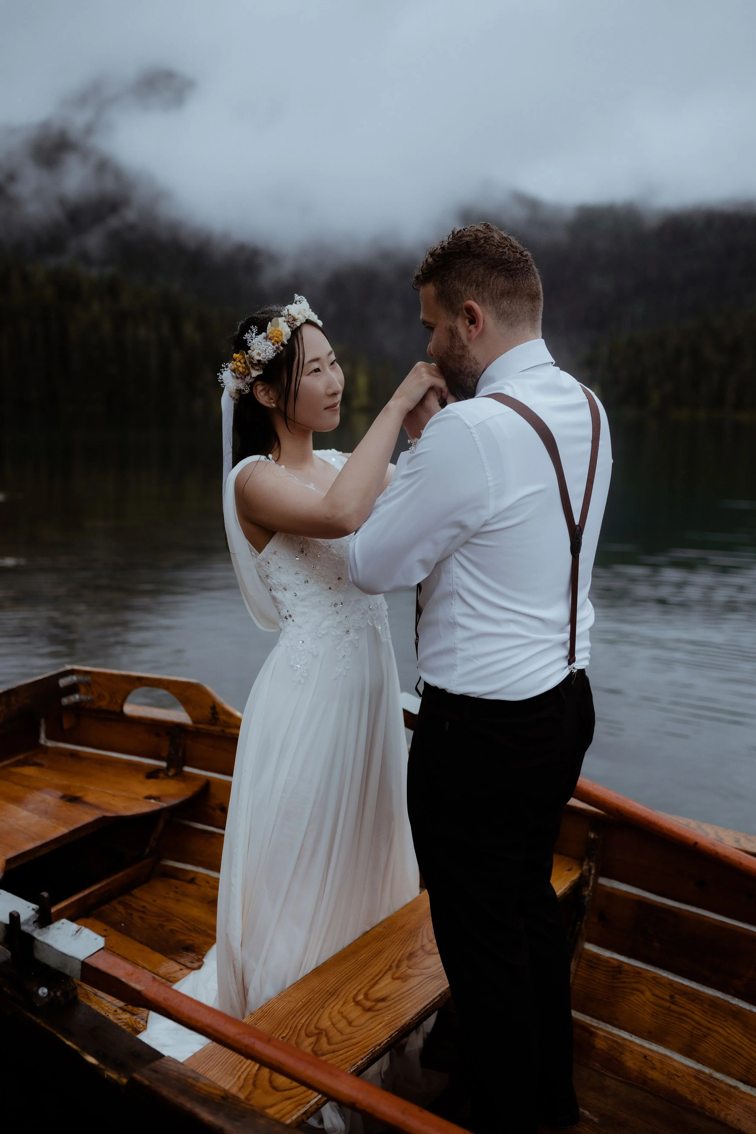 A groom and bride sharing a romantic moment on a wooden boat, with a misty lake and mountains in the background.