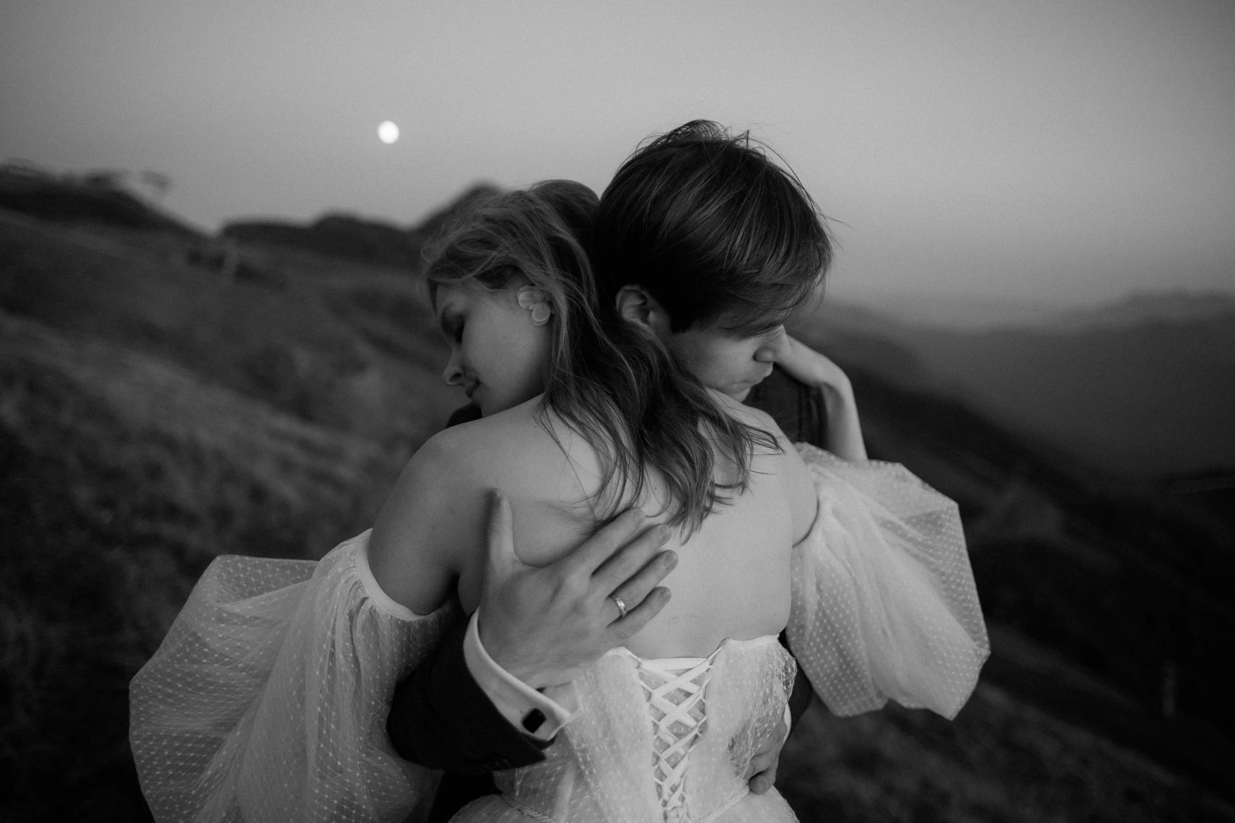 Black and white photo of a couple embracing outdoors at dusk, with a full moon in the background.