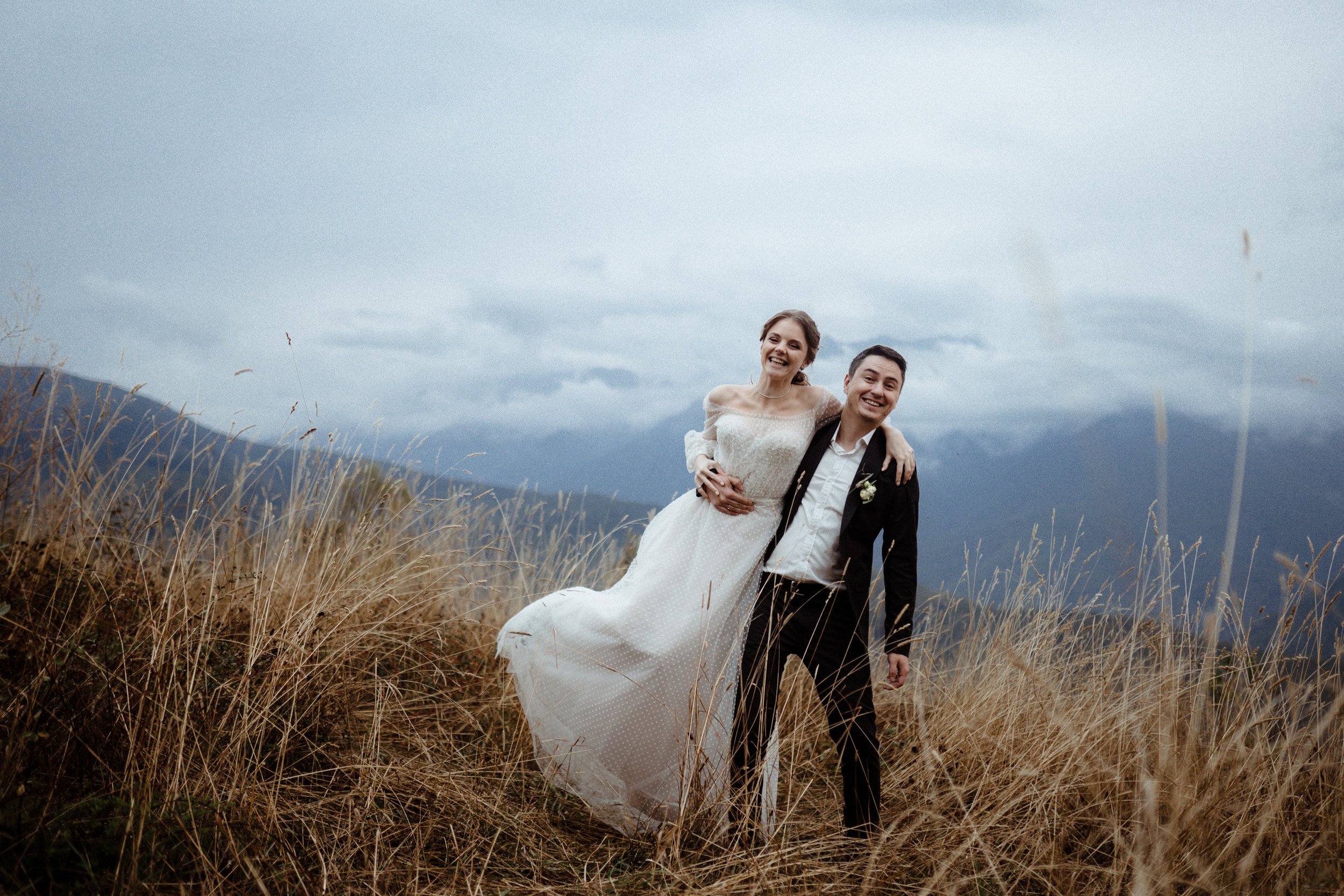 A smiling woman in a wedding dress being carried by a man in a suit, standing in tall grass outdoors with mountains and a cloudy sky in the background.
