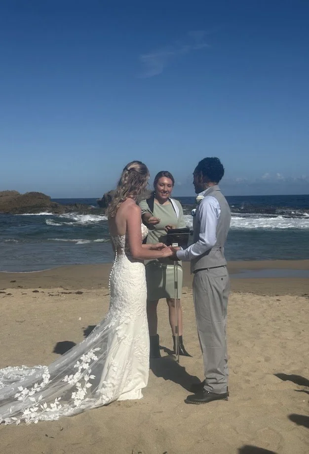 Paradise Vows PR founder Kendra marrying a couple on the beach. The bride is in a long, flowing white gown and the groom is in a grey suit. 