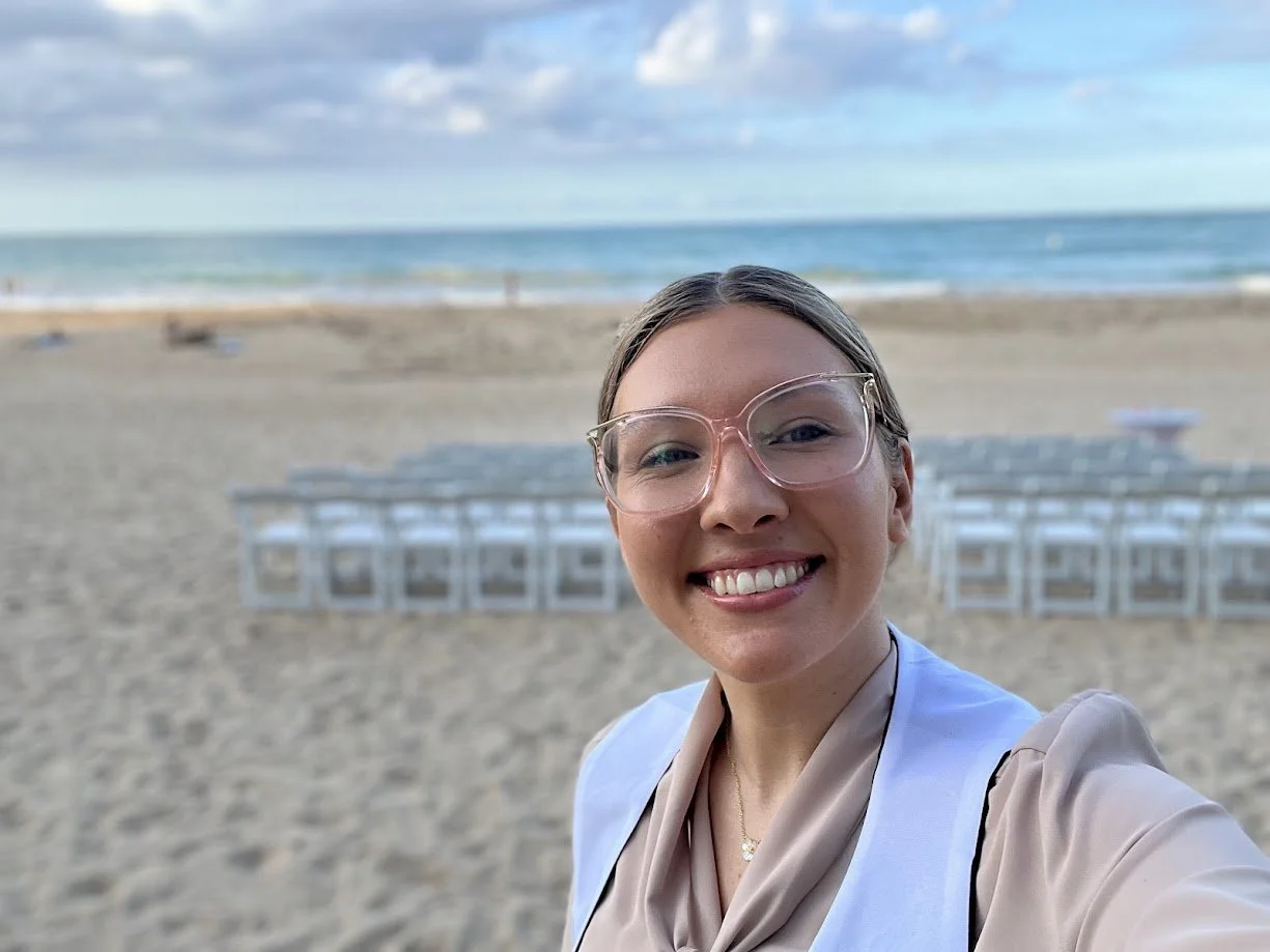 Paradise Vows PR founder Kendra smiling on the beach with a destination wedding set up in the background looking about the ocean. 