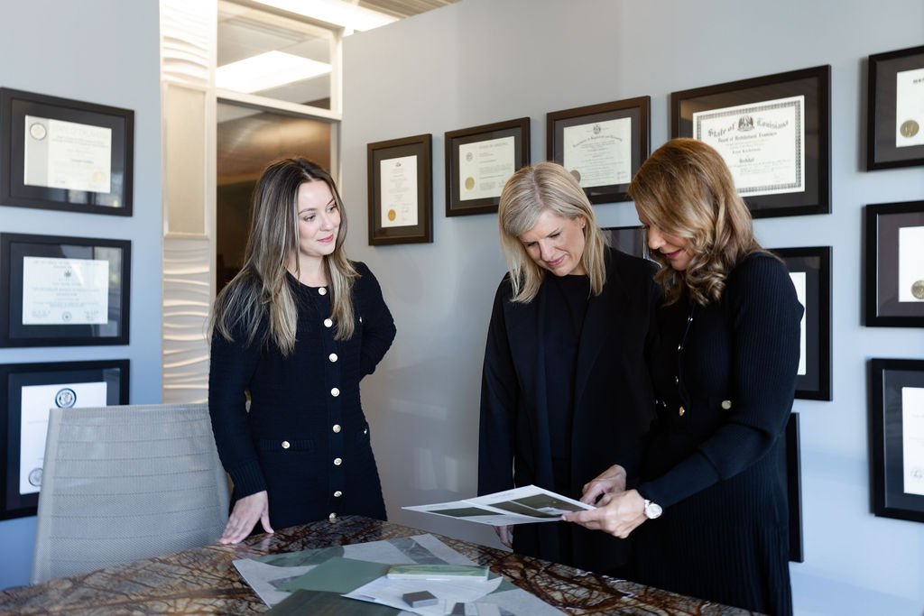 Three women review documents in a conference room with framed certificates on the wall.