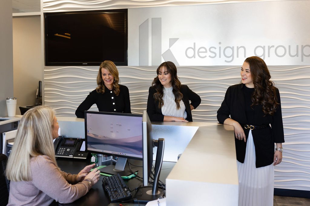A woman sitting at a reception desk using a computer, talking to three women standing in front of her, inside an office with a sign reading 'K design group' on the wall.