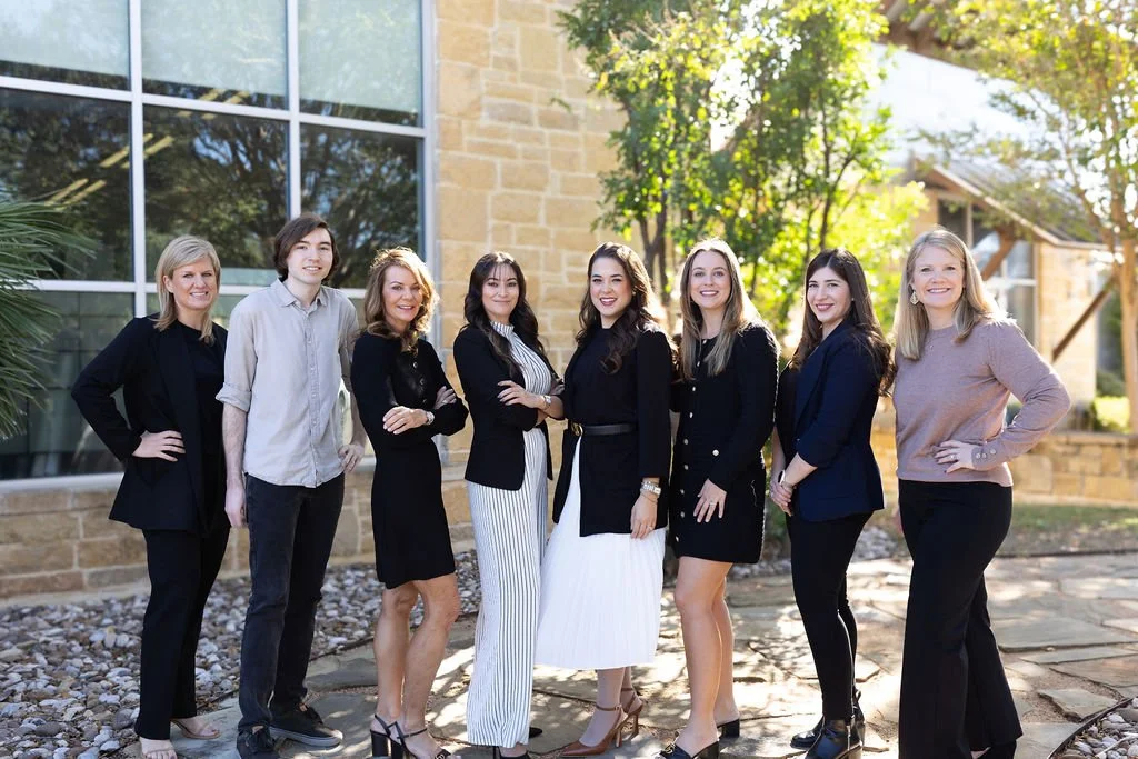 Group of seven women standing outdoors in front of a building with large windows and trees, dressed in business casual attire, smiling at the camera.