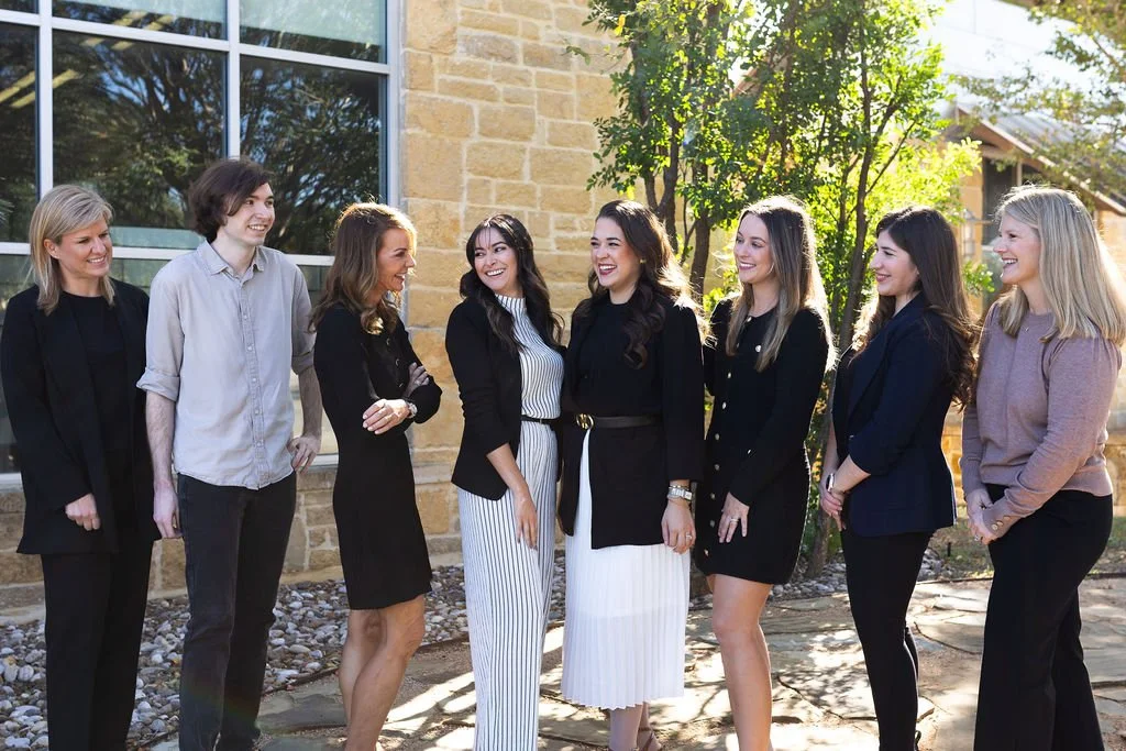 Group of eight women standing outdoors, smiling and talking, with a building and trees in the background.