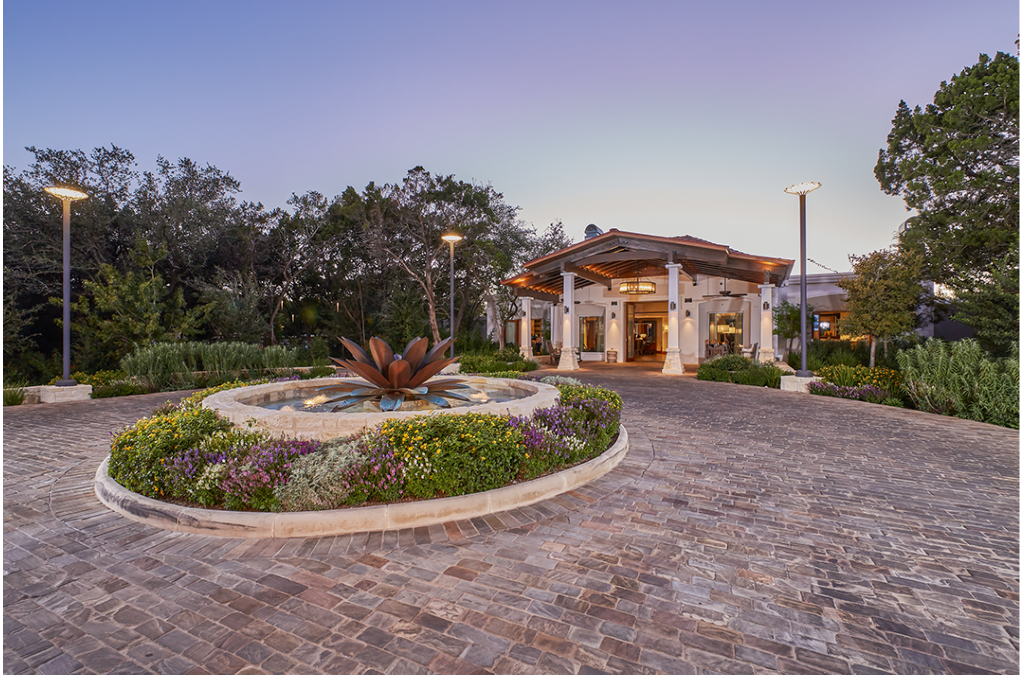 A private estate with a paved courtyard, a decorative fountain surrounded by flowers, and a white house with a wooden roof and outdoor seating.