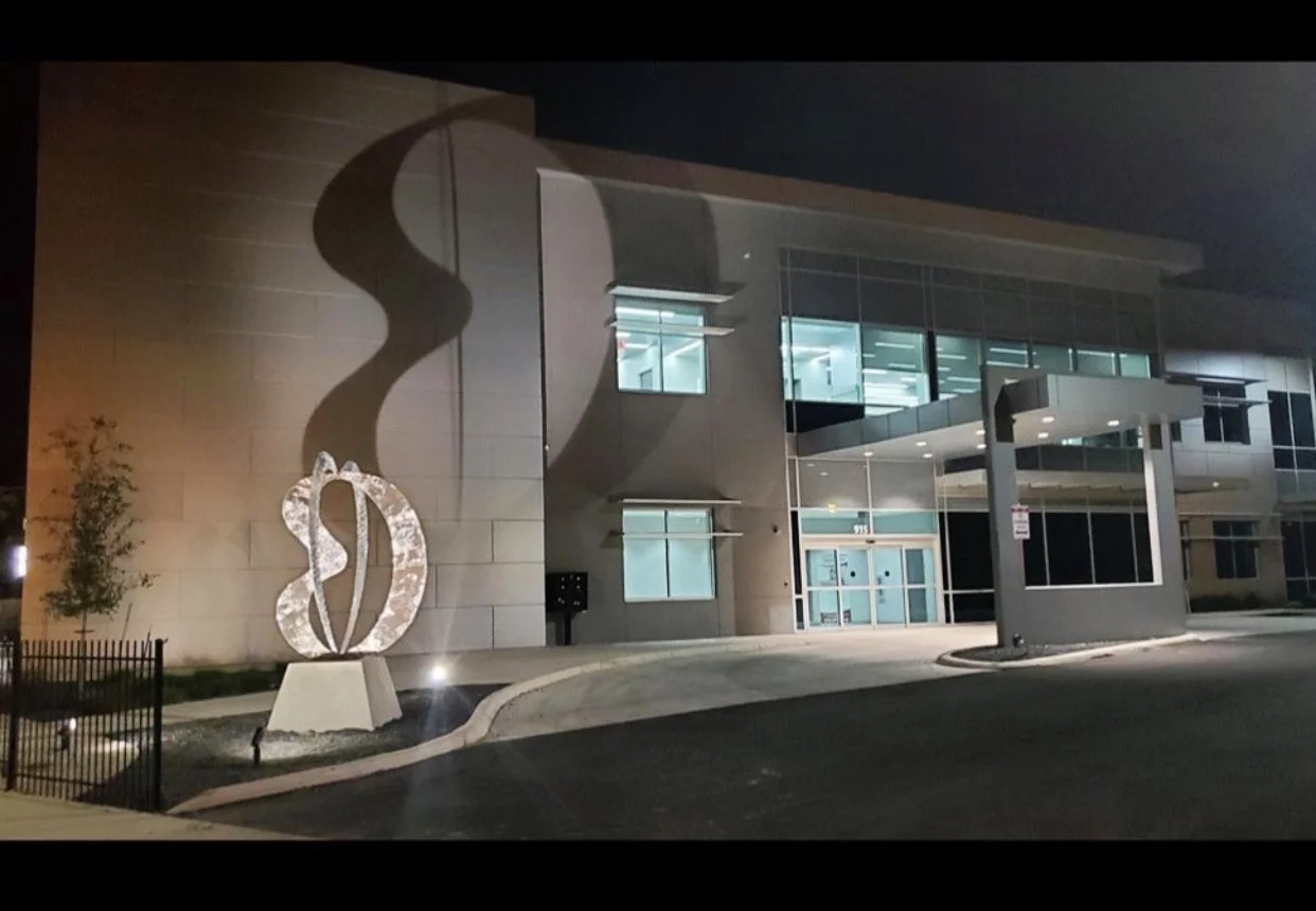 Night view of a modern building with large glass windows and a large abstract sculpture of a musical note outside, casting a shadow on the wall.