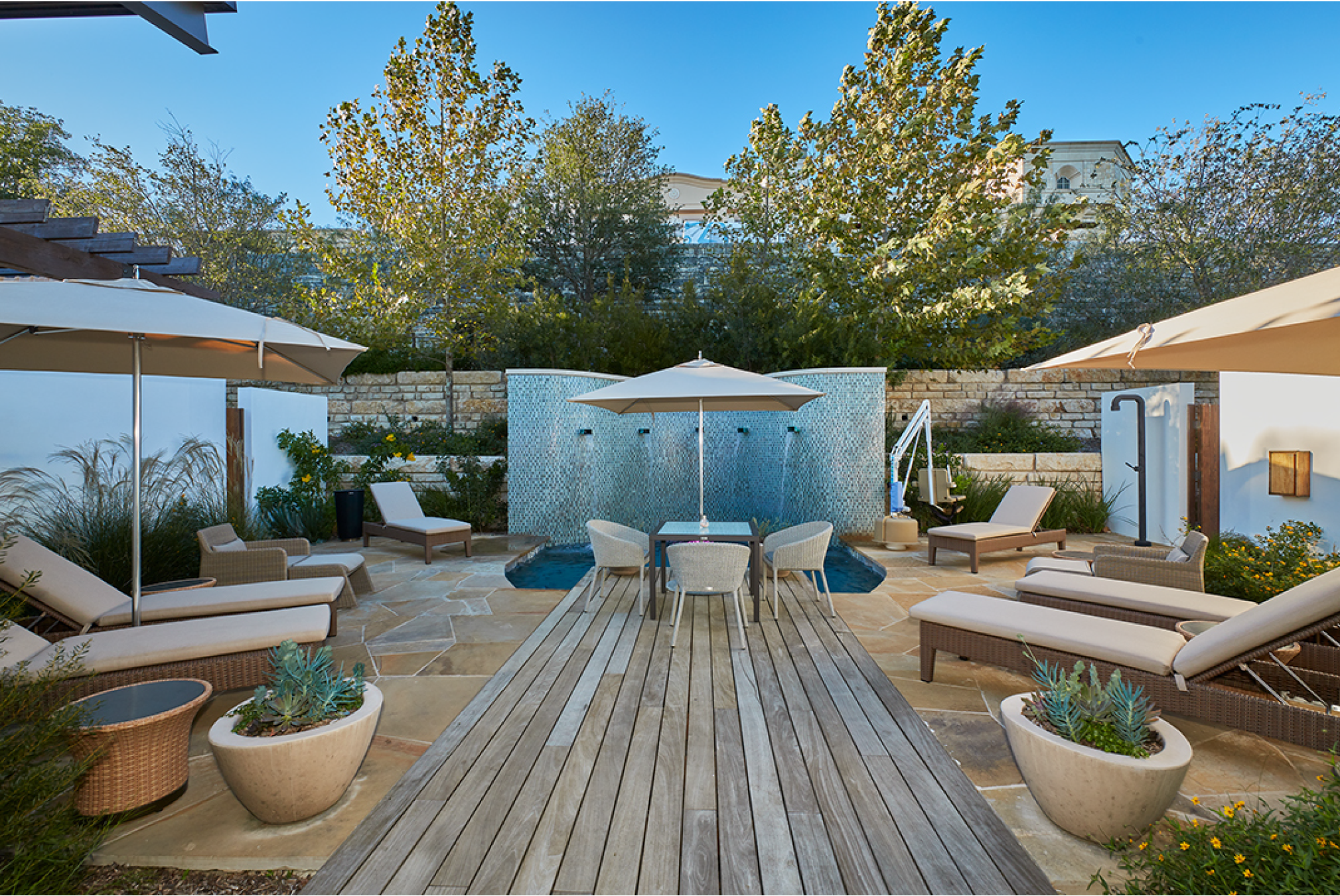 Outdoor pool area with lounge chairs, umbrellas, small tables, plants, and a central table with chairs, surrounded by a stone patio and trees.