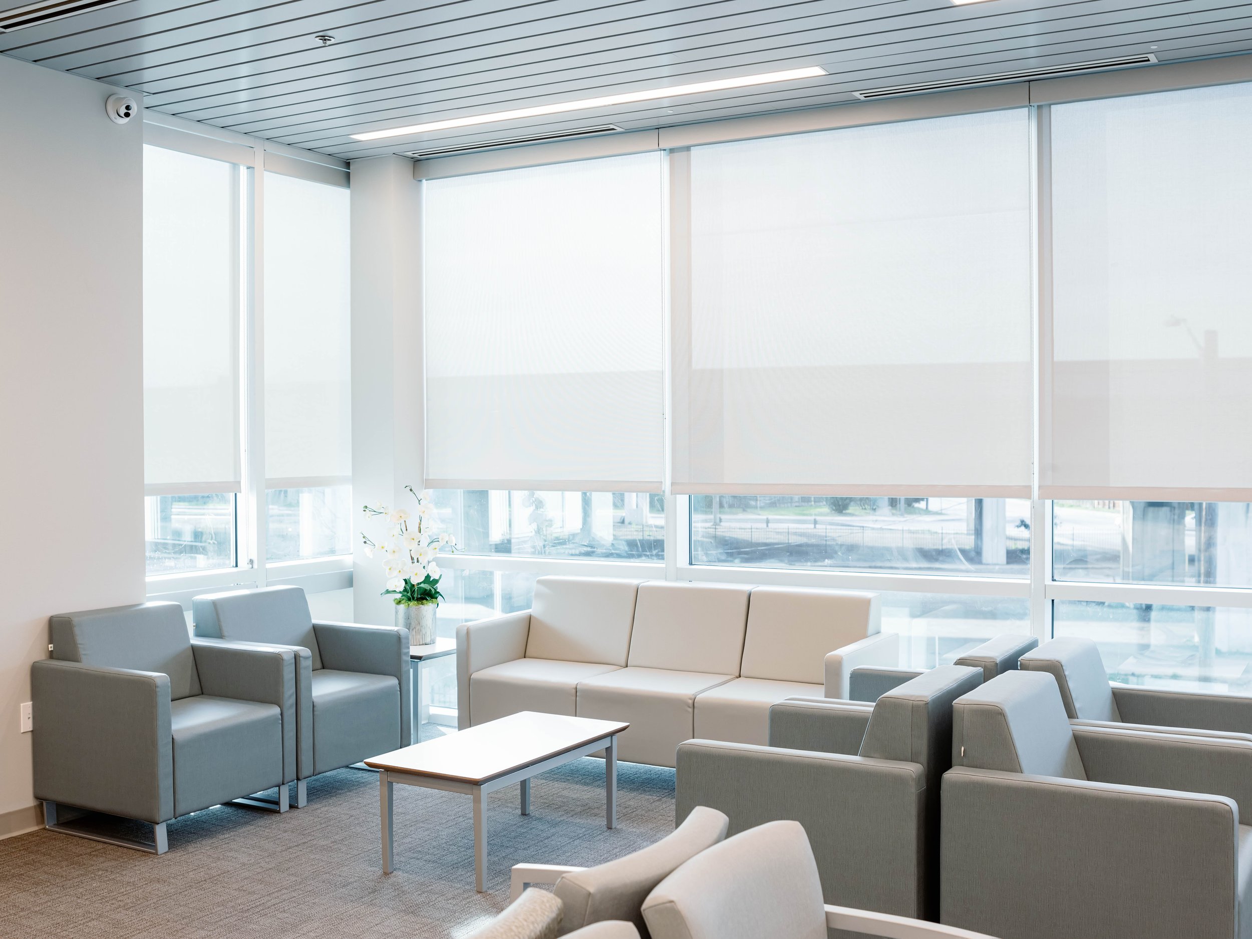 Modern airport lounge with gray armchairs, a white sofa, small tables, large windows with blinds, and a potted orchid plant.