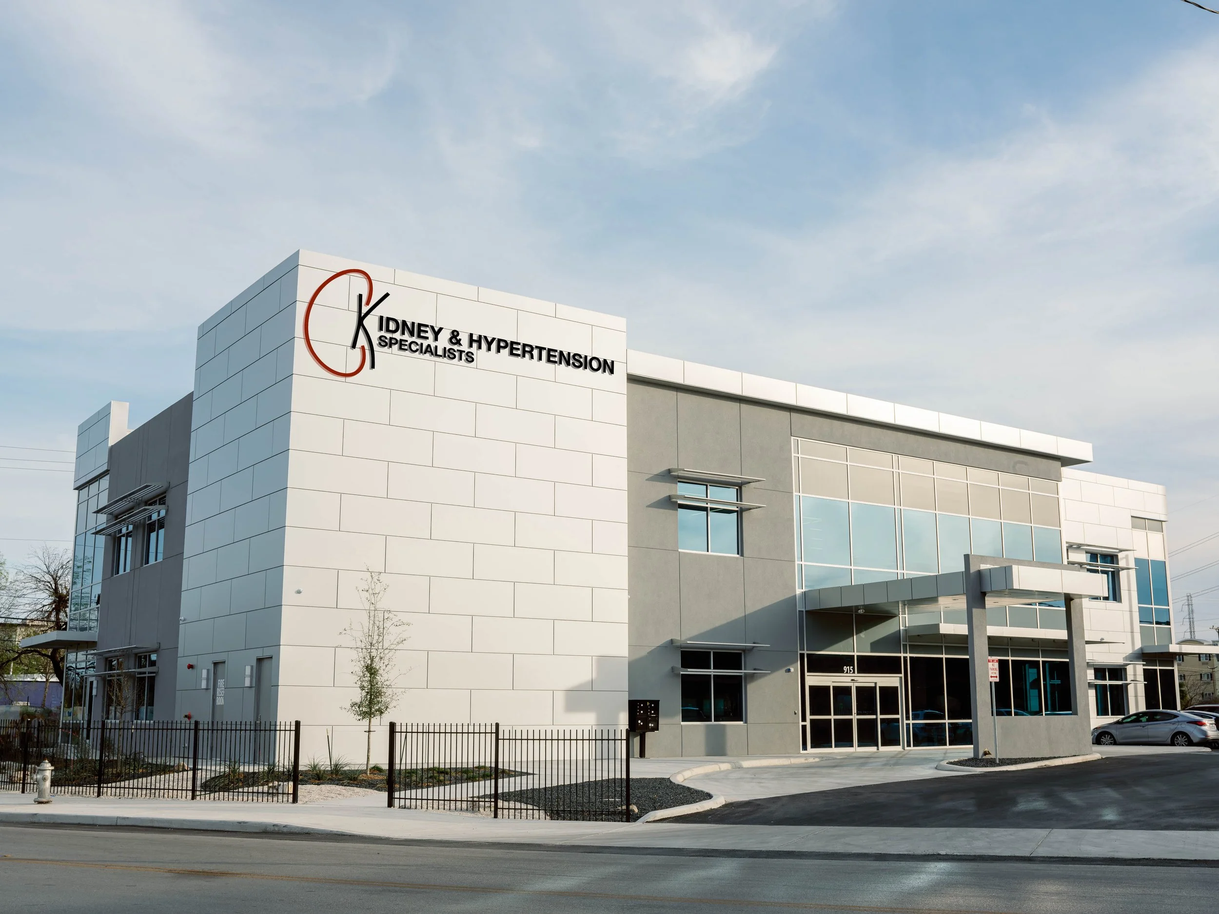 Modern medical clinic building with sign that reads Kidney & Hypertension Specialists, large windows, and a small fenced landscaping area in the foreground.