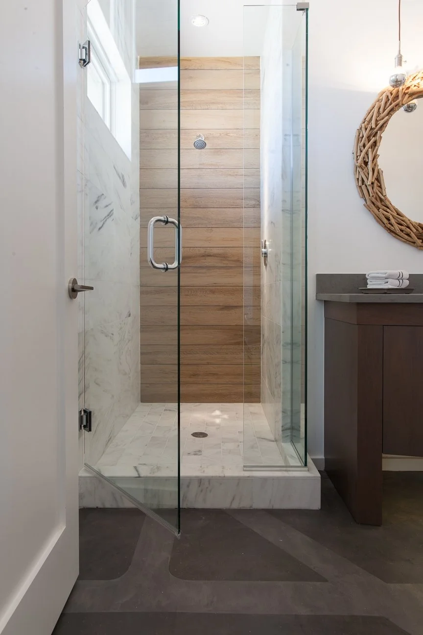 A modern glass shower stall with marble flooring and wood-paneled wall inside, next to a white vanity with rolled towels and a round woven mirror on the wall.