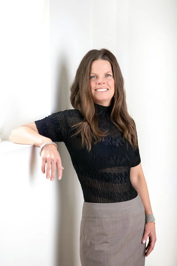 A woman with long brown hair wearing a black short-sleeve top and beige skirt, standing against a white wall, smiling softly.
