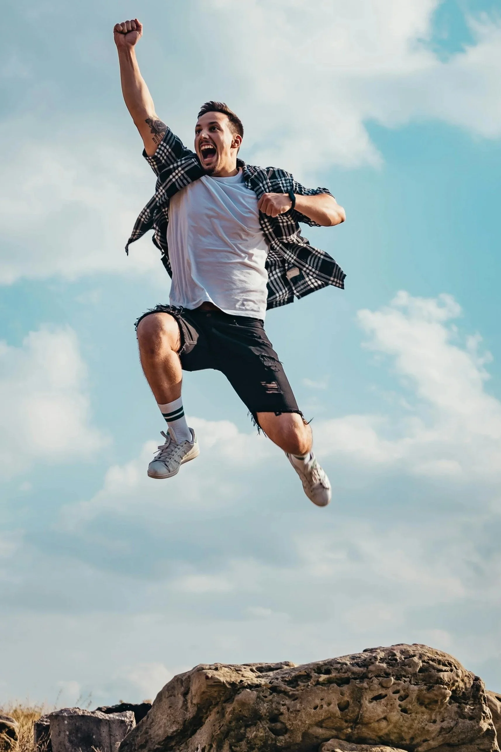 A man bouncing joyfully in mid-air above rocks outdoors with a cloudy sky background, wearing a white t-shirt, black shorts, sneakers, and a plaid shirt.