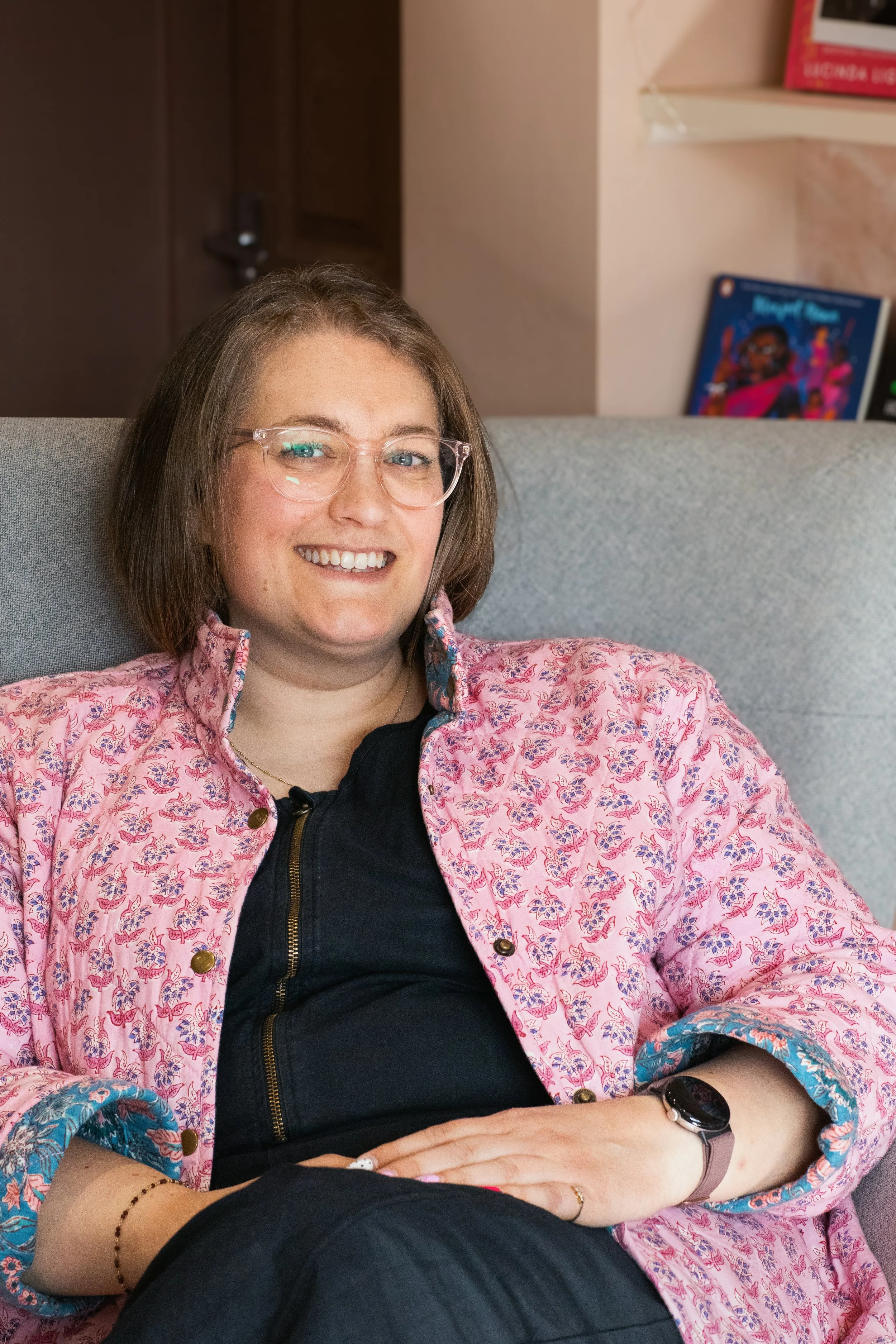 A woman with shoulder-length brown hair, glasses, and a big smile, sitting on a gray couch wearing a pink floral shirt over a black top, with a watch on her left wrist and a bracelet and ring on her right hand, in a cozy room with books and artwork in the background.