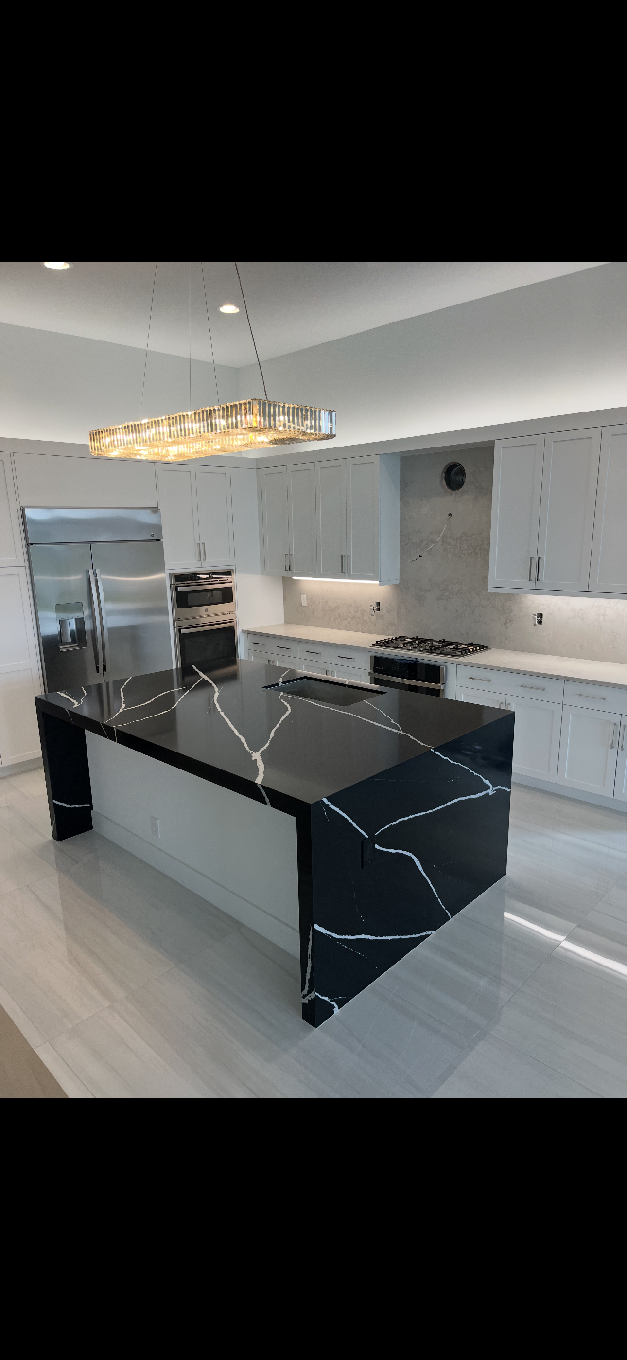 Modern kitchen with a black marble-topped island, white cabinetry, stainless steel refrigerator, and a built-in oven. The ceiling has recessed lighting and a rectangular crystal chandelier.